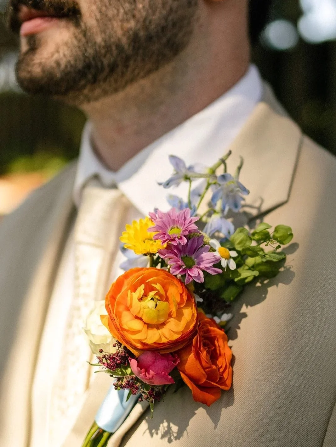 Close-up of a man's lapel with a colorful flower boutonniere attached, featuring orange, pink, purple, yellow, white, and green flowers, on a beige suit at a formal event.