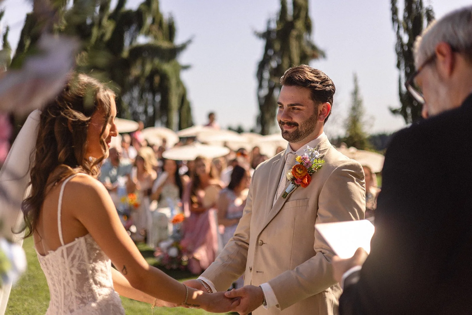A couple getting married outdoors, holding hands and exchanging vows, with guests and umbrellas in the background on a sunny day.
