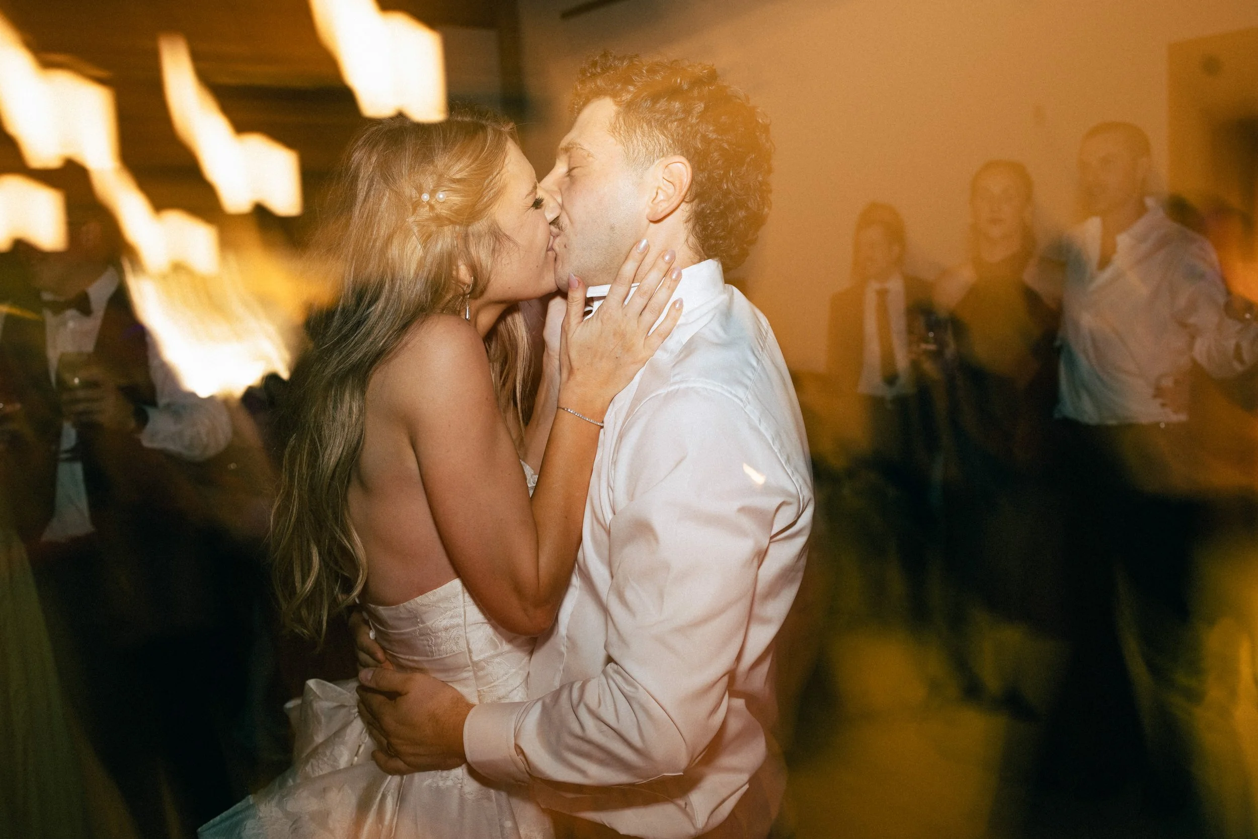 A couple sharing a kiss at a celebration or wedding, with people laughing and enjoying in the background.