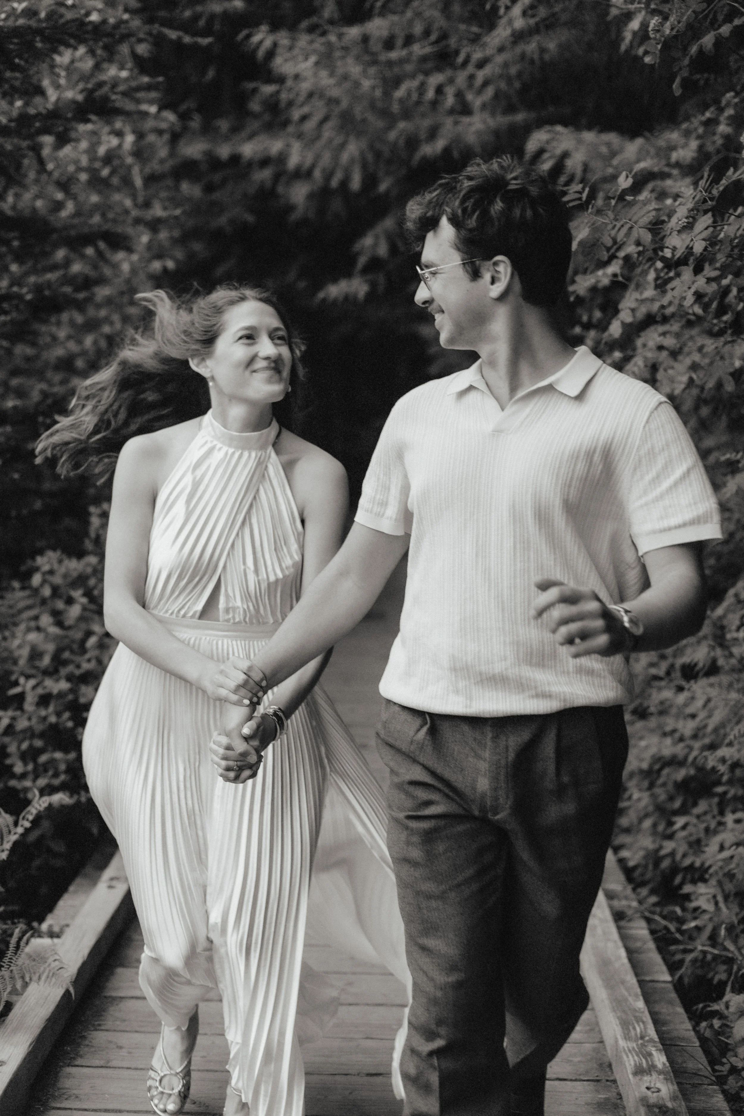 A black and white photo of a couple walking hand in hand outdoors. The woman is smiling and looking at the man, while the man is looking at her, both smiling. They are walking on a wooden bridge with foliage in the background.