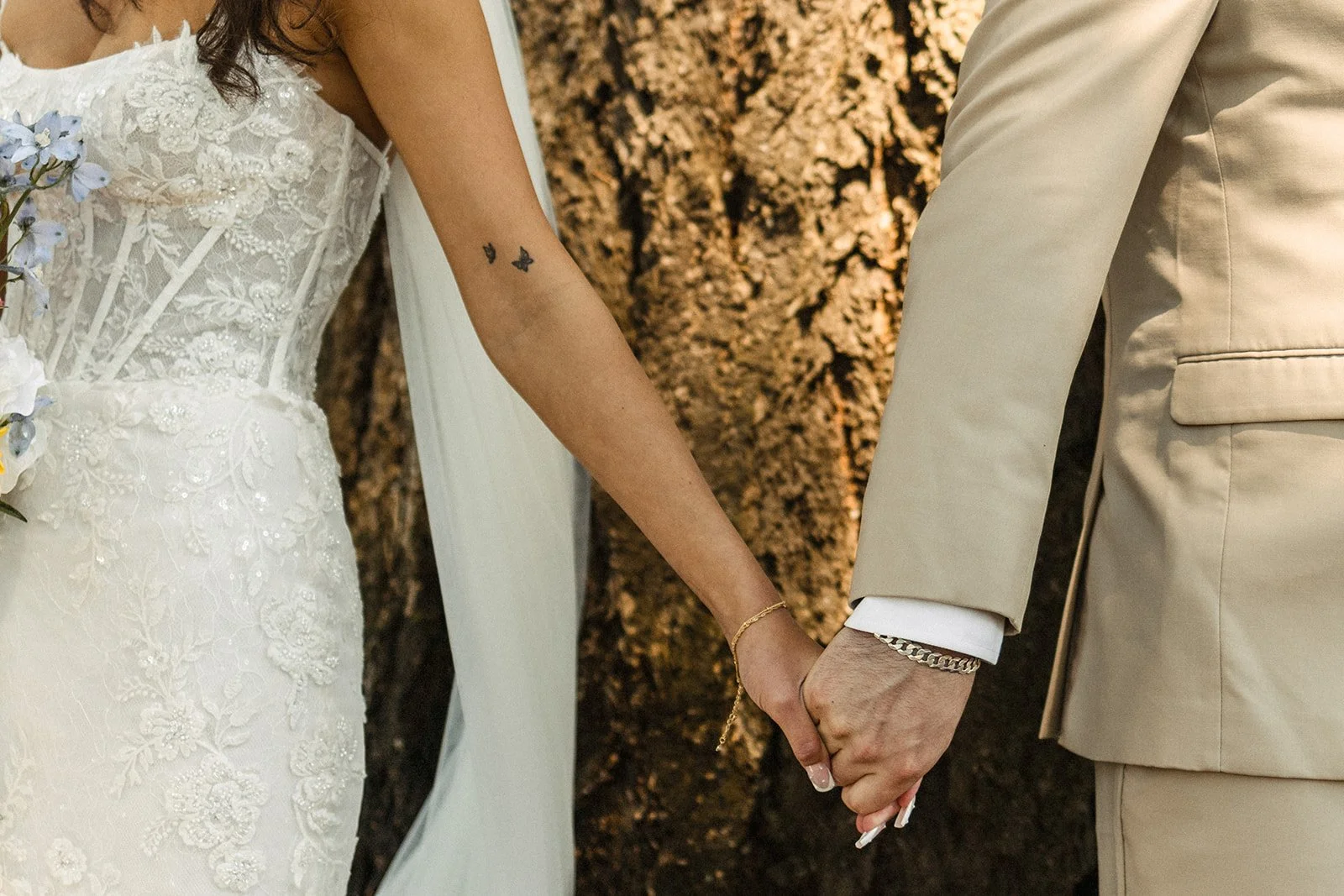 A bride and groom holding hands during their wedding, with the bride wearing a white lace wedding dress and the groom wearing a beige suit, set against a background of trees.