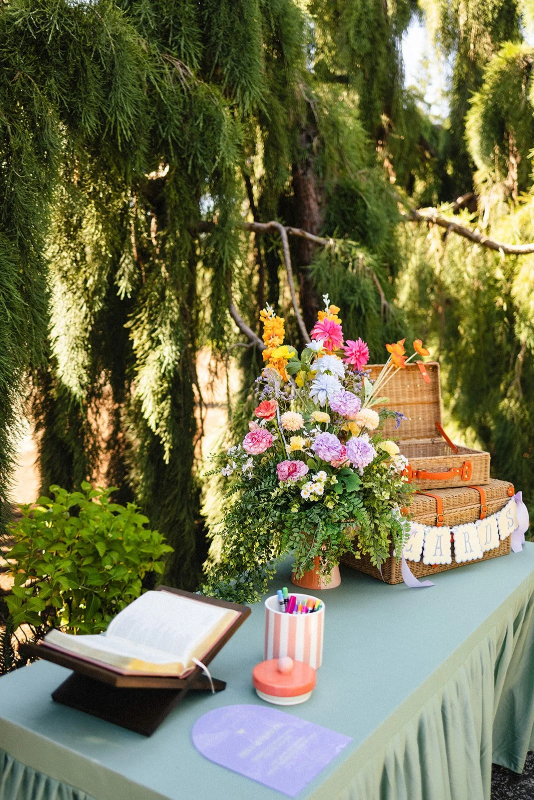 A table decorated for a celebration with a large floral arrangement, an open book, a container of colorful pens, and a wicker picnic basket, with a sign that reads 'HAPPY BRIDAL SHOWER' hanging on the basket, set outdoors surrounded by lush green tre