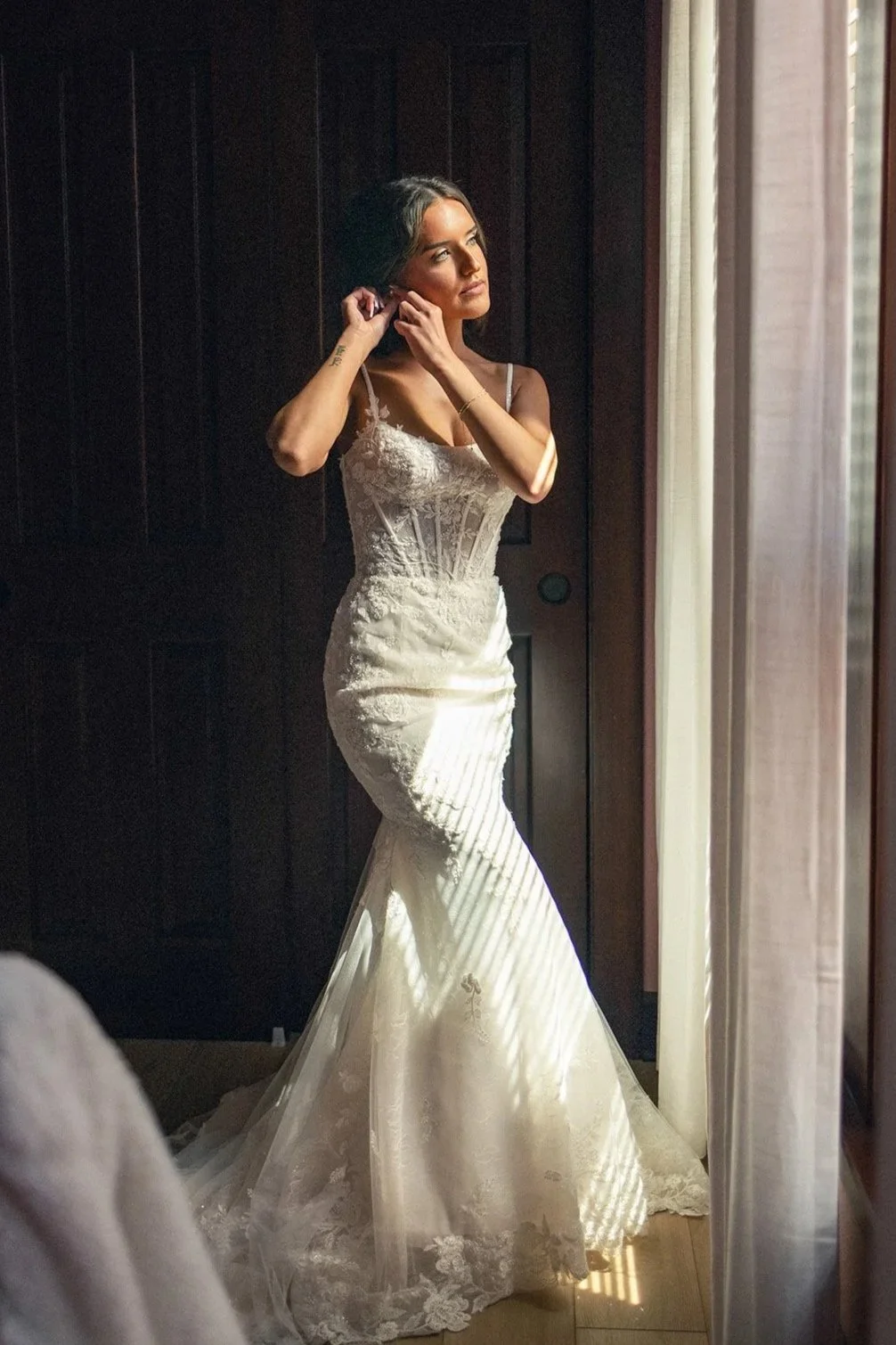 A woman in a white wedding dress standing by a window, adjusting her earring and looking thoughtfully out of the window, with sunlight streaming in and creating shadows on the floor and her dress.
