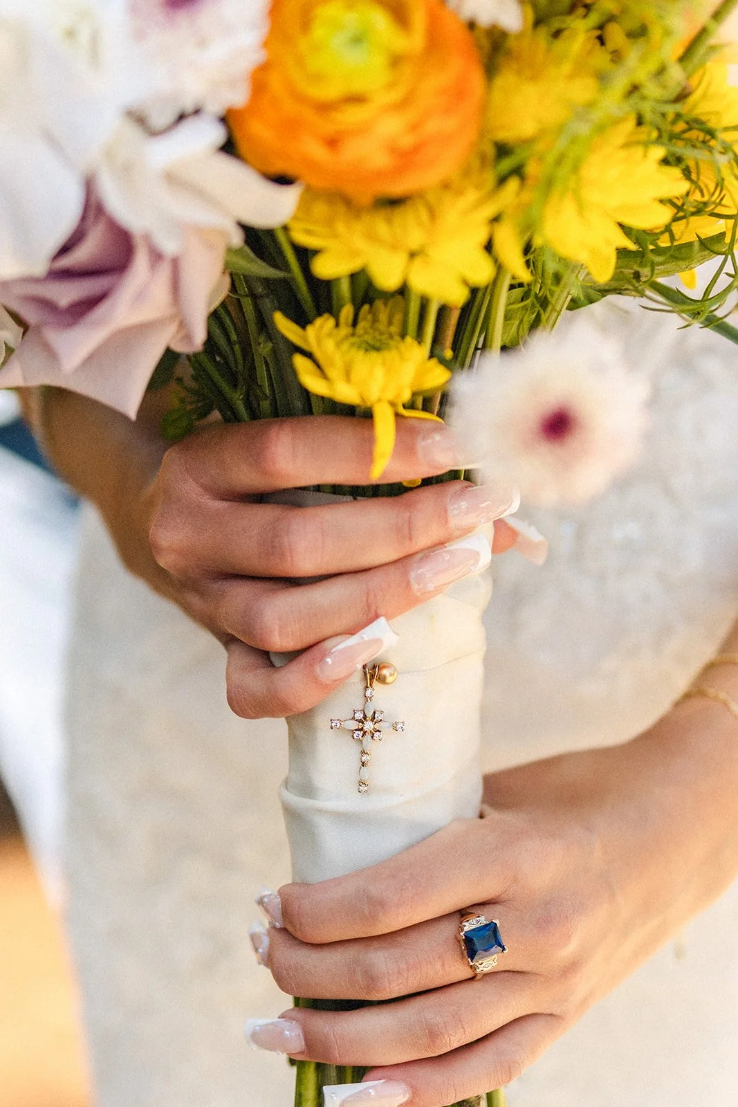 Close-up of a woman's hand holding a bouquet of yellow, white, and orange flowers, with a cross and pearl ring on her finger, and a blue gemstone ring on her other hand.