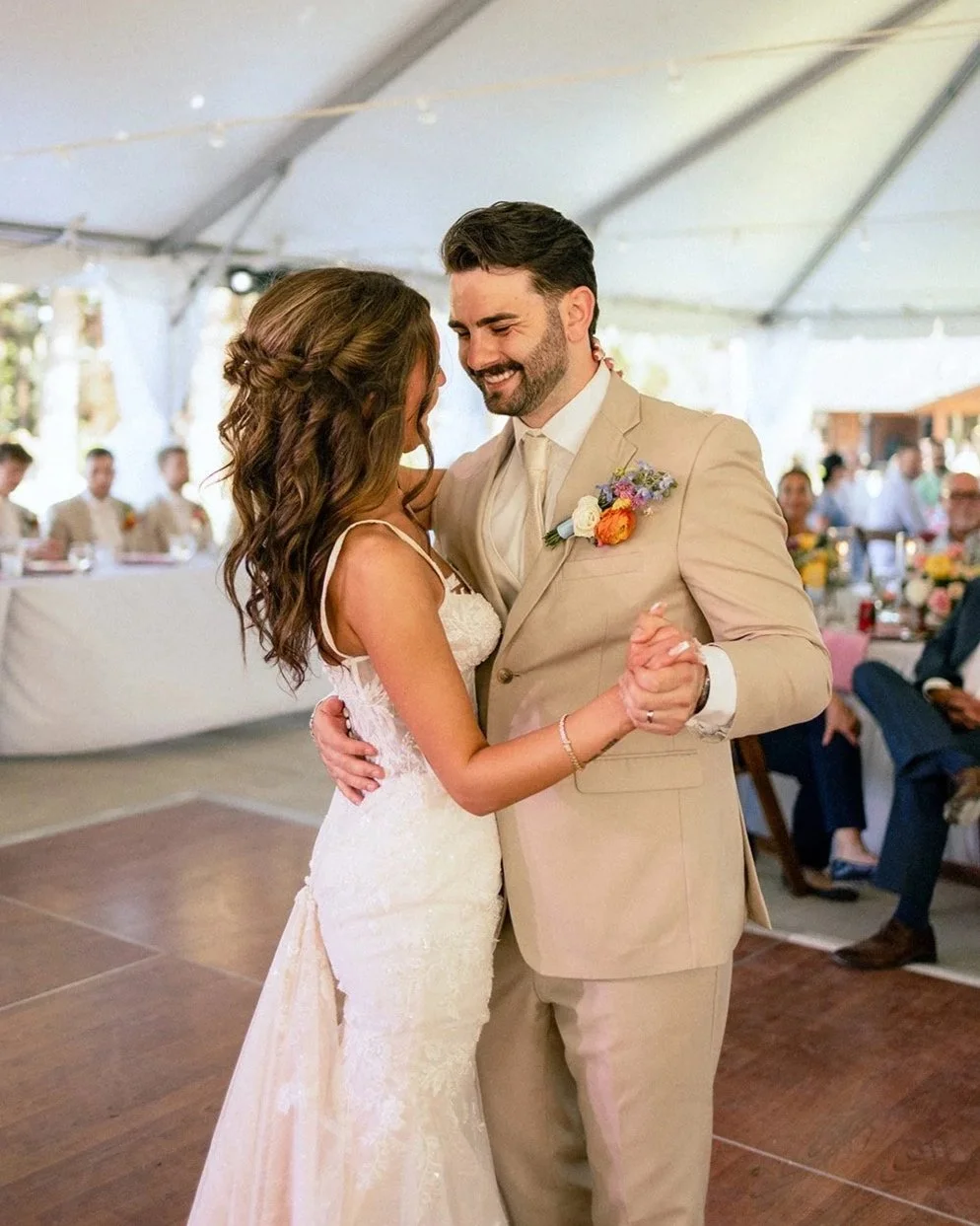 A bride and groom dancing at their wedding reception inside a large tent, surrounded by seated guests. The bride wears a white lace wedding dress, and the groom wears a beige suit with a boutonniere. They are happily dancing and smiling.