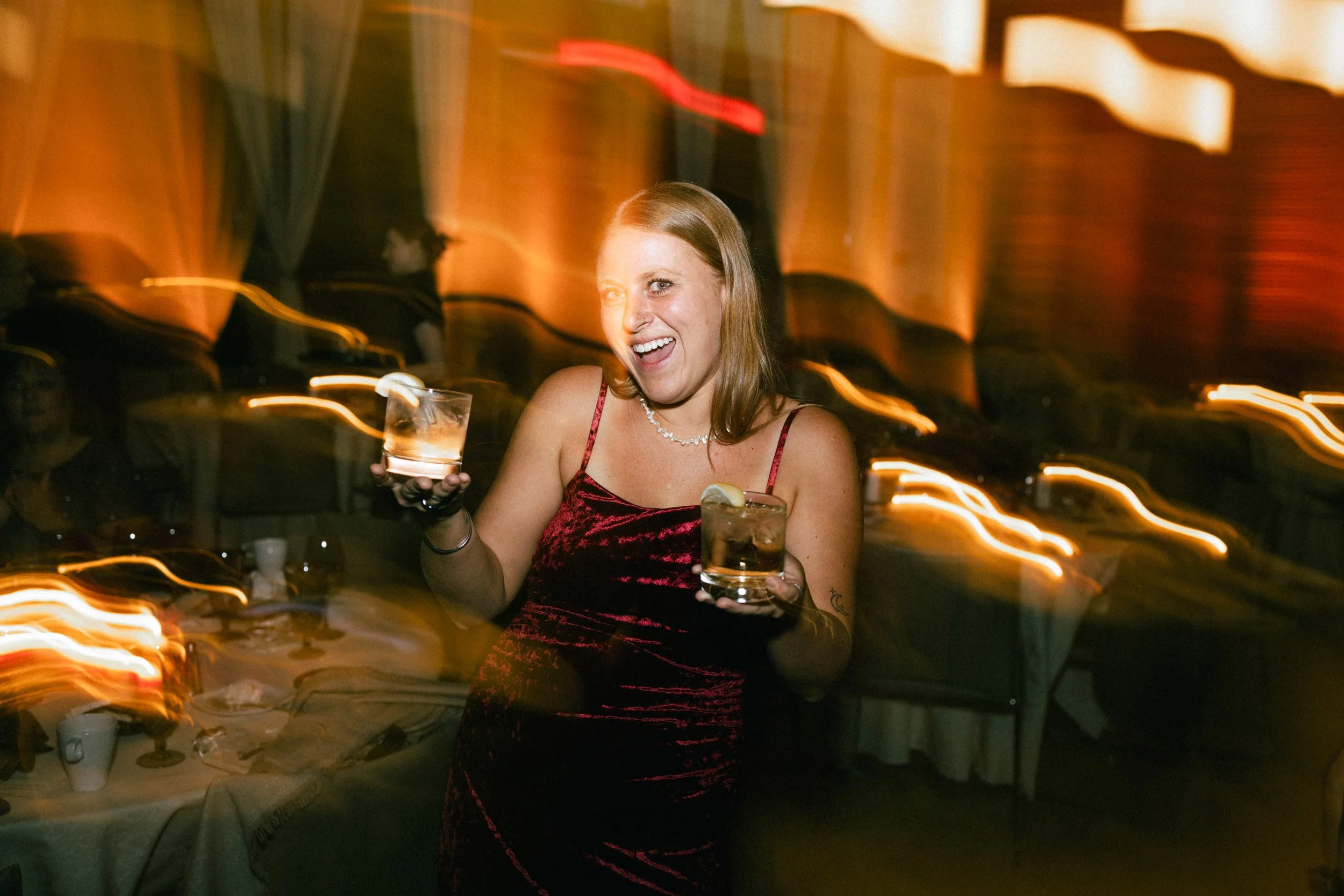 A woman in a red velvet dress holding a drink with lemon slice in a dimly lit party or celebration setting.