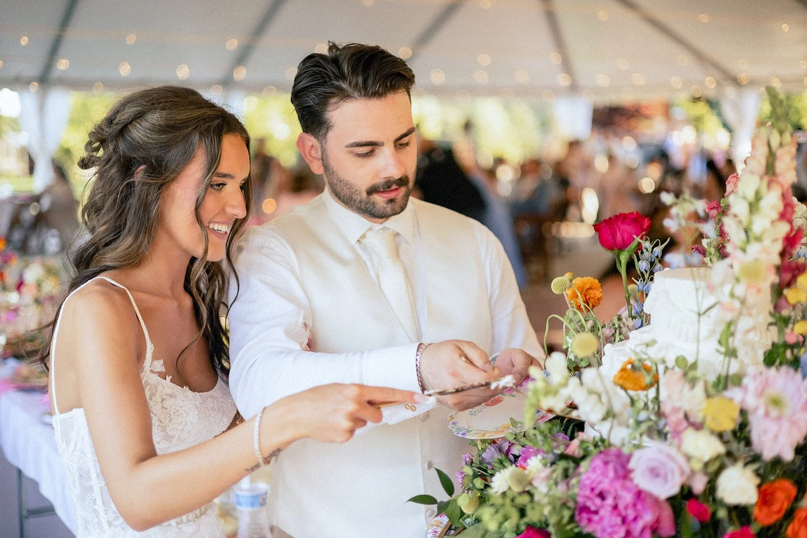 A bride and groom cutting their wedding cake under a decorated tent with colorful flowers.