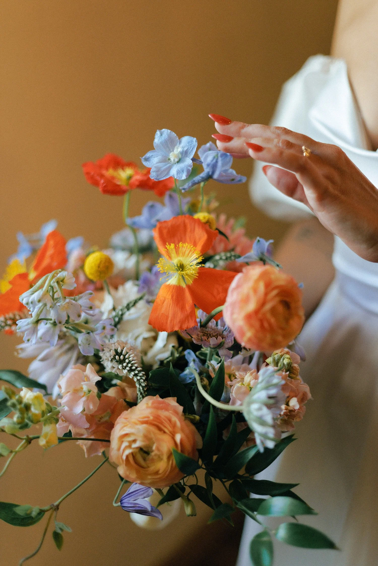 A close-up of a woman's hand with painted red nails reaching towards a vibrant, colorful bouquet of flowers against a neutral background.