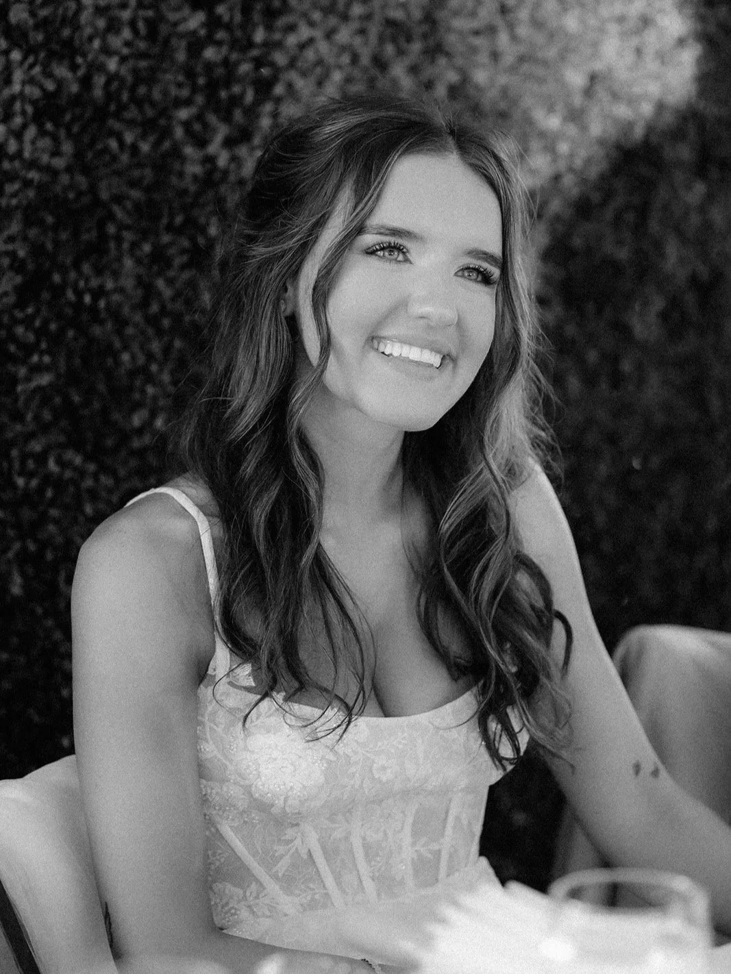 A woman with long wavy hair and a big smile, wearing a sleeveless dress, sitting at a table with a blurred glass in the foreground, outdoors at night.