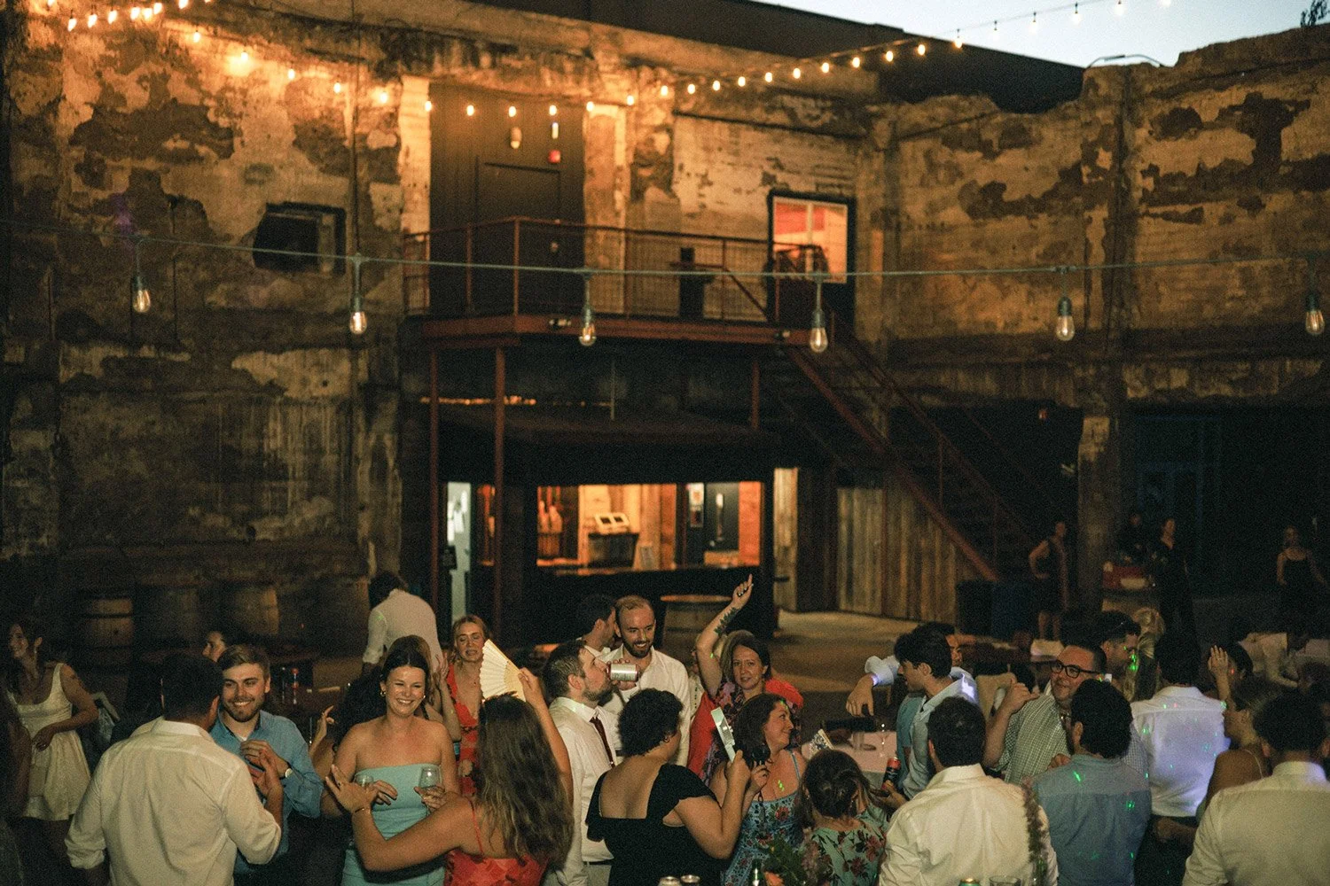 Group of people dancing and socializing at an indoor party in a rustic, old warehouse with exposed brick walls, string lights, and a small stage area.