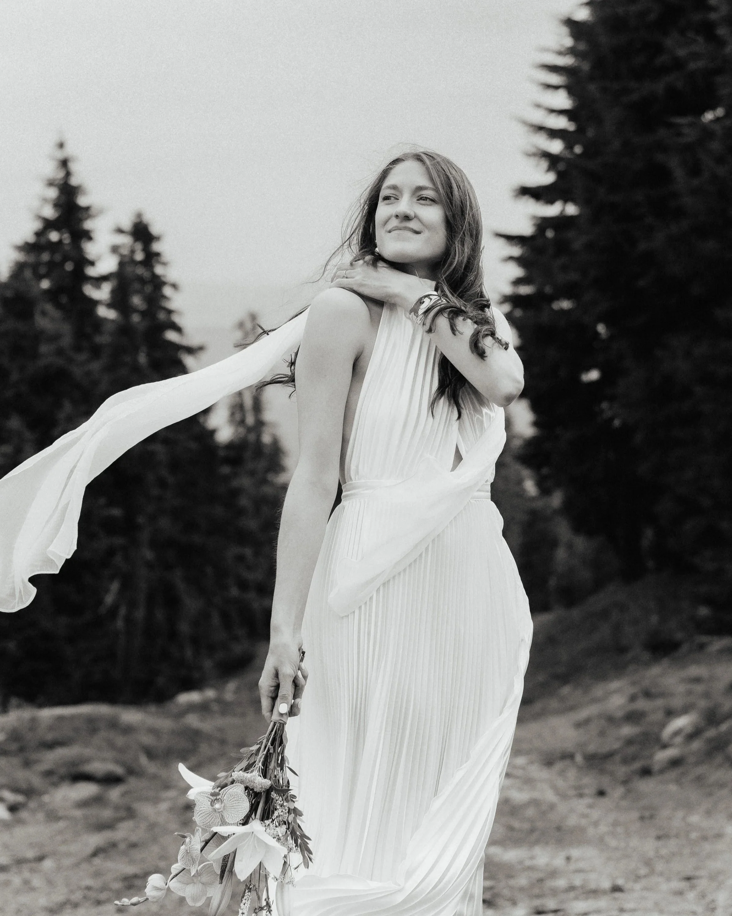A woman in a white dress holding a bouquet of flowers standing outdoors with pine trees in the background. The image is in black and white.