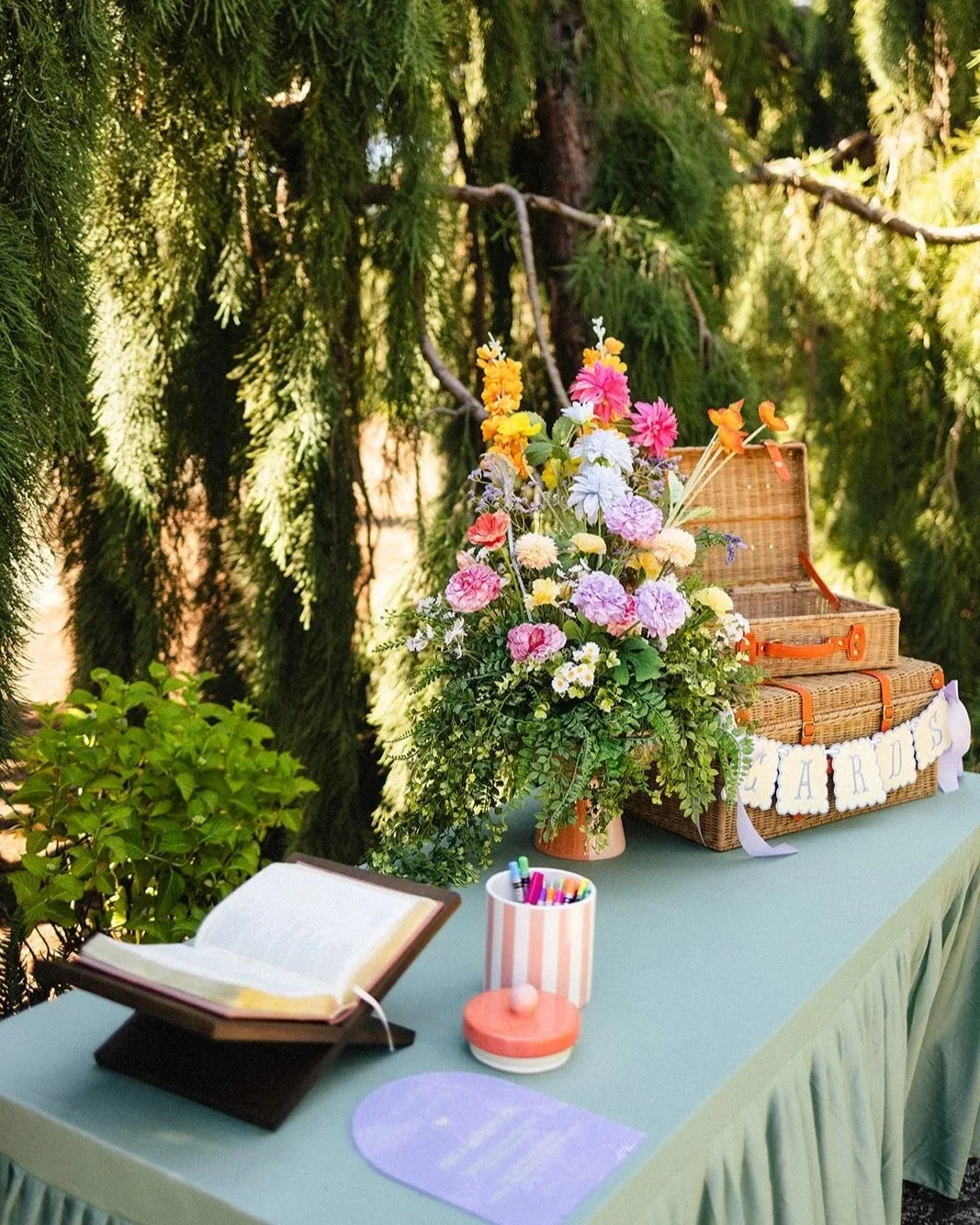 A table decorated with a colorful flower arrangement and a vintage suitcase with a 'Cards' banner, outdoors among greenery, with markers, an open book, and a small pink and white container on the table.
