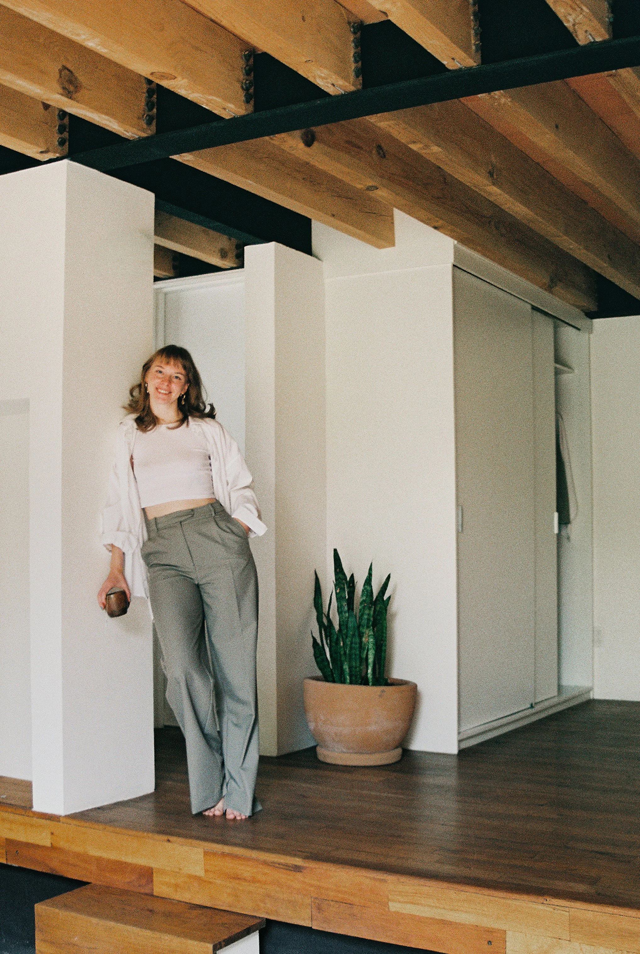 Woman smiling and standing on a wooden stage with a potted plant beside her, in a room with white walls and wooden ceiling beams.