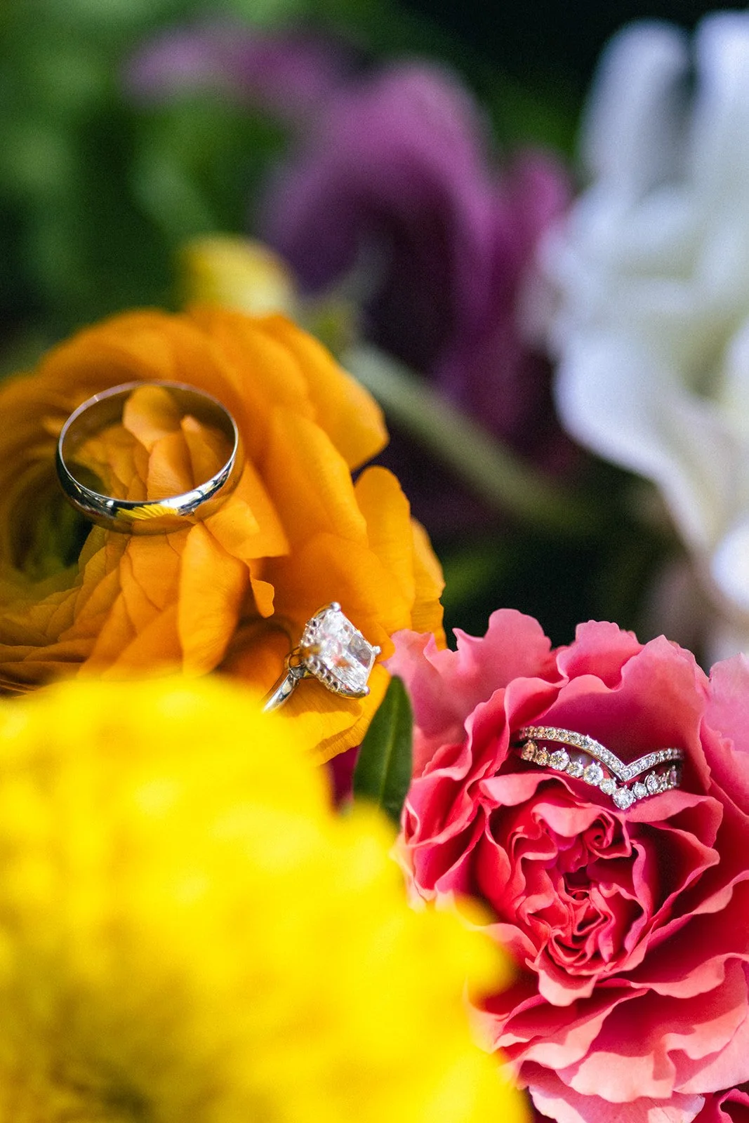 Close-up of a bouquet of colorful flowers with two rings resting on the petals, one with a large center stone and the other a band with smaller stones.