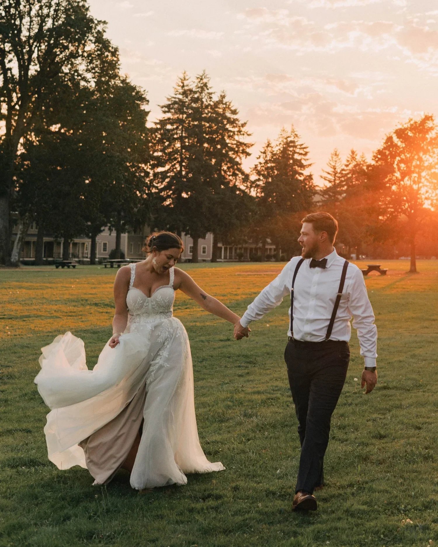 A bride and groom hold hands, smiling as they walk across a grassy field at sunset during their Portland wedding.