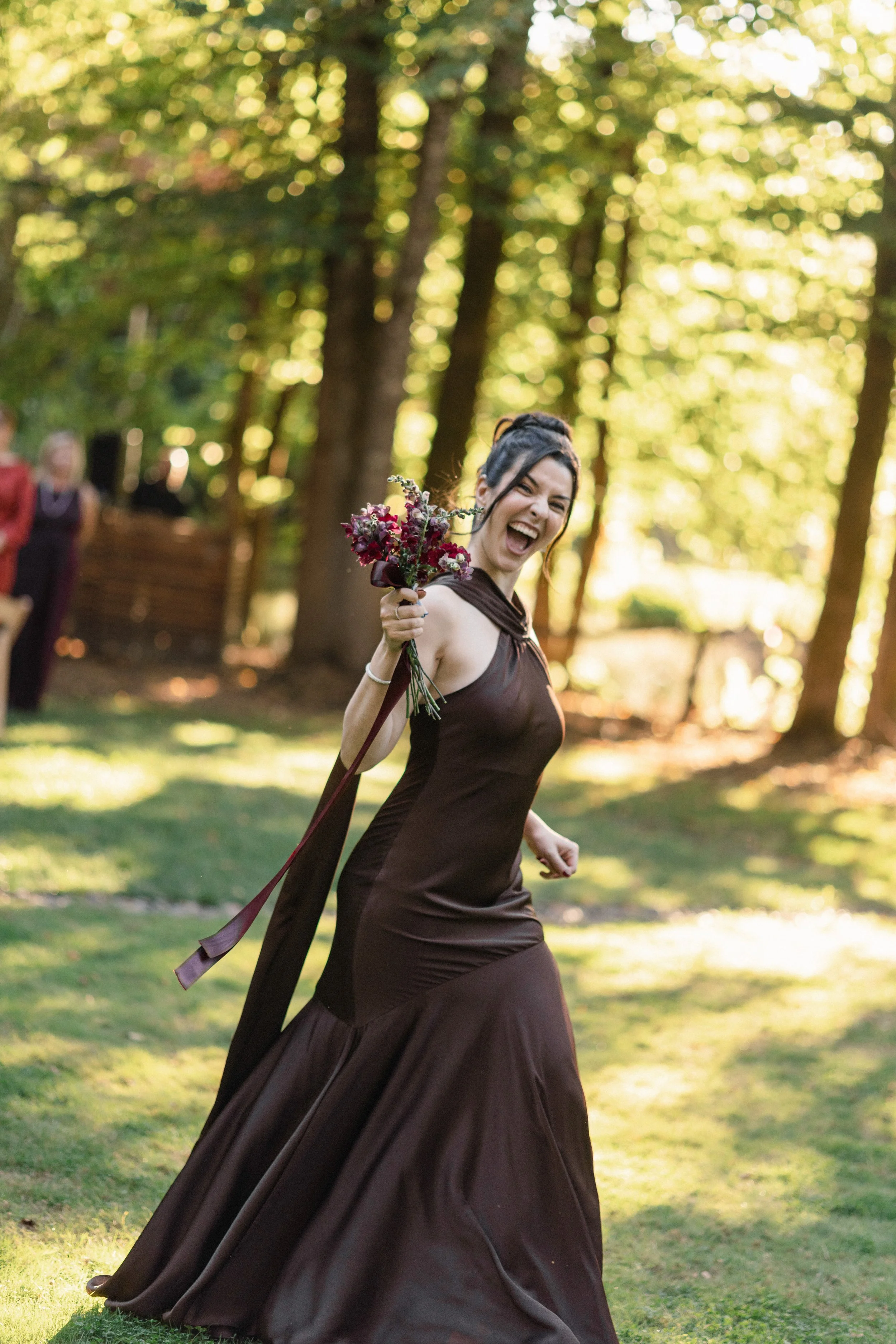 Woman in a brown dress holding a bouquet of flowers, smiling and dancing in a park with sunlight filtering through trees.