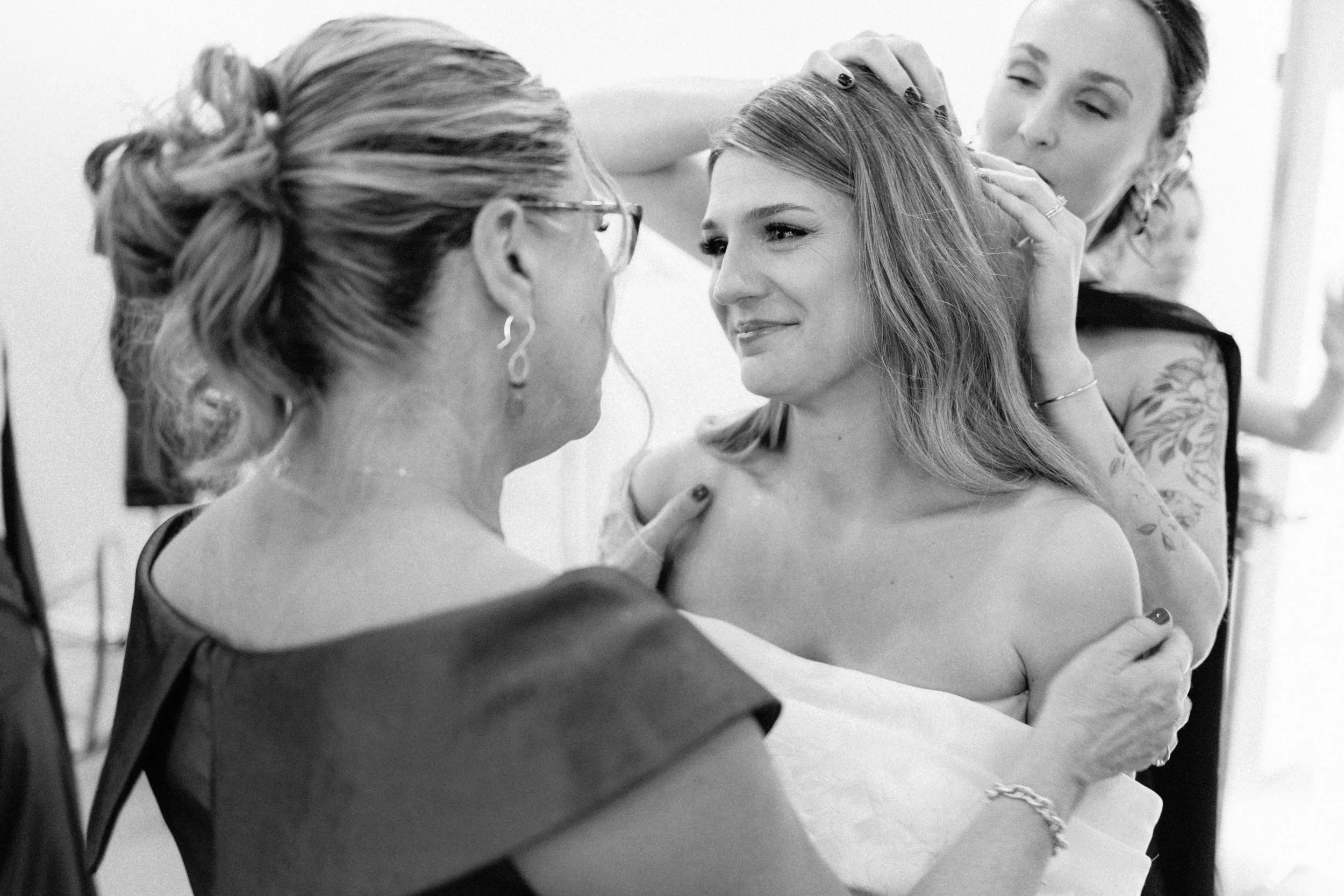 A woman with long hair is being attended to by two women, likely during a wedding or special event. The woman in the center is smiling and looking at one of the women assisting her, while the other woman is fixing her hair. The scene appears to be intimate and cheerful.