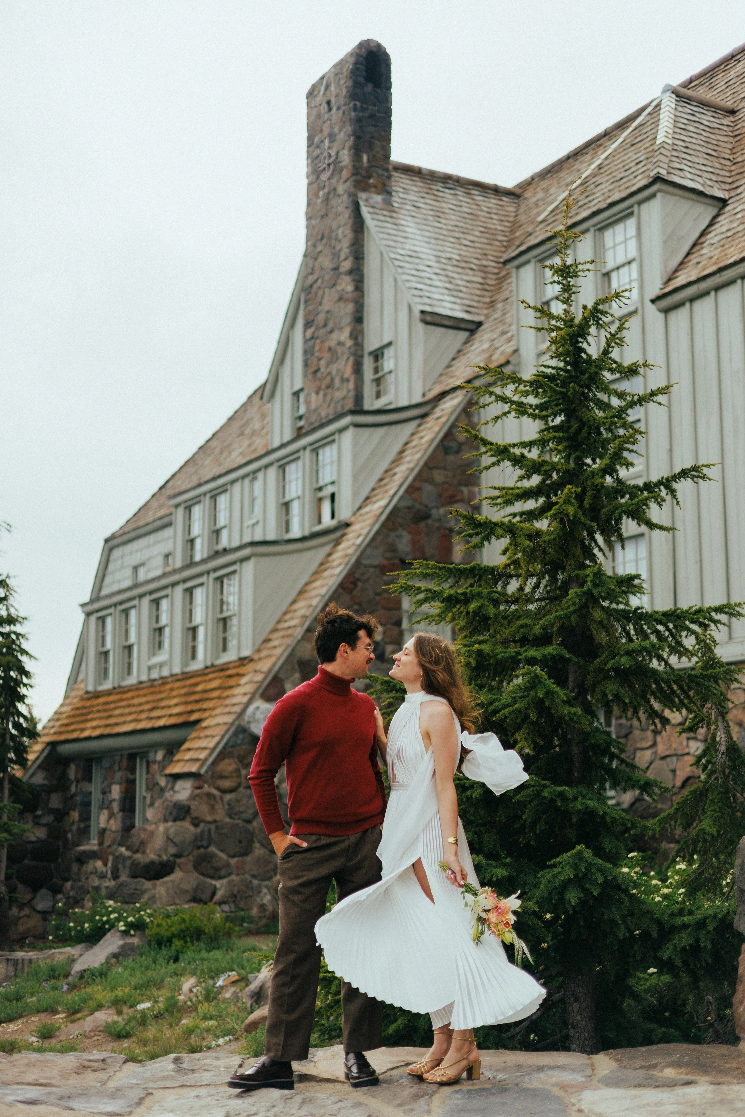 A couple stands outdoors in front of a large house with a steep roof and stone chimney, surrounded by greenery. The woman wears a white dress and holds a bouquet, while the man wears a red sweater and brown pants.
