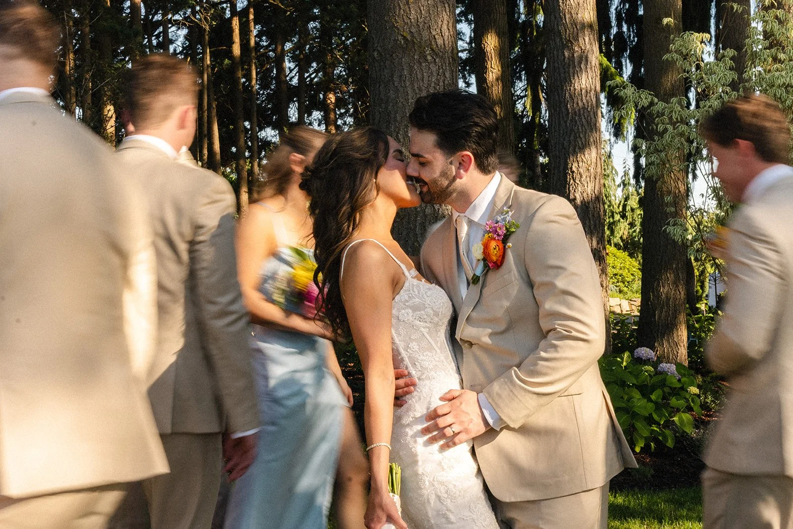 A bride and groom are kissing during their wedding ceremony outdoors, with friends or family blurry around them in a wooded area.