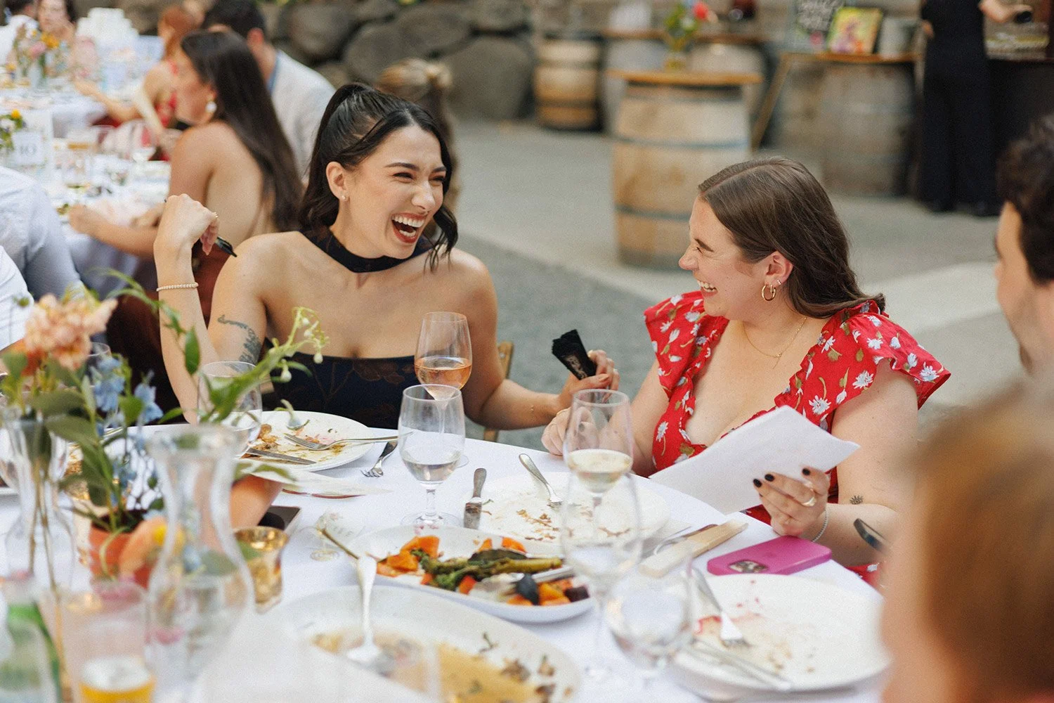 Two women laughing and enjoying themselves at a dinner party, seated at a table with food and drinks.