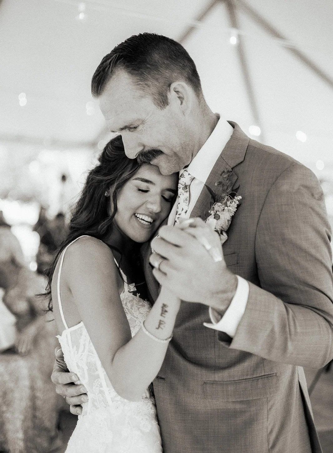 A black and white photo of a bride and groom dancing together, embracing lovingly with eyes closed, smiling. The bride has long dark hair and wears a lace wedding dress, the groom has short dark hair, a mustache, and is dressed in a suit with a boutonniere.