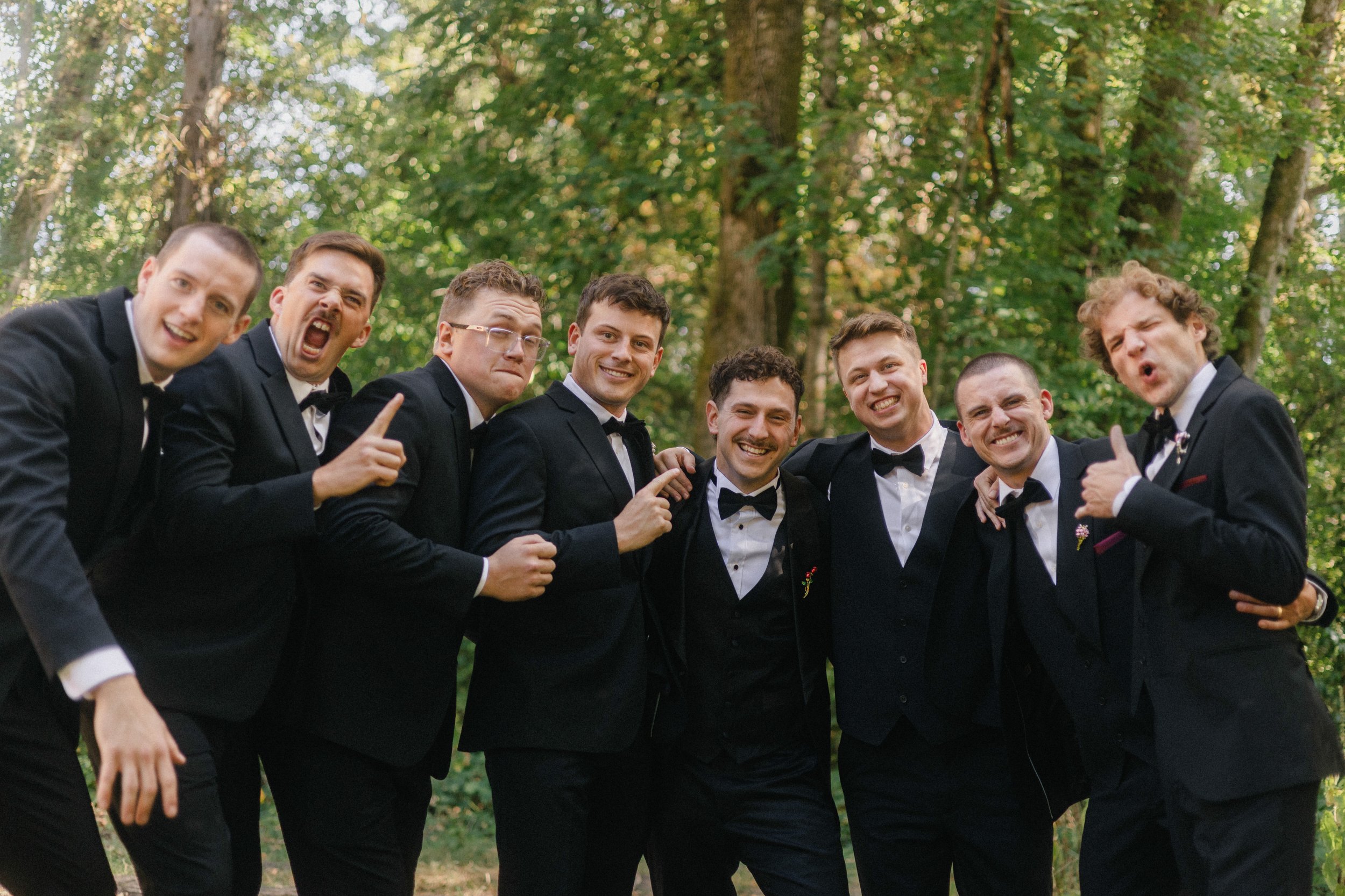 Group of nine men dressed in tuxedos smiling and posing outdoors in a wooded area.
