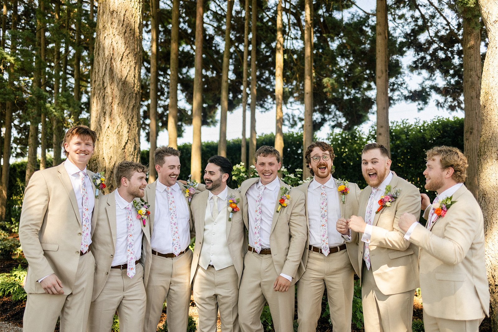 A group of nine men dressed in beige suits with floral ties, standing outdoors among trees at a wedding, smiling and laughing.