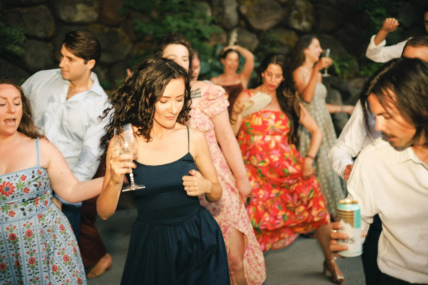 People dancing and socializing outdoors at a party, some holding drinks, surrounded by rocks and greenery.