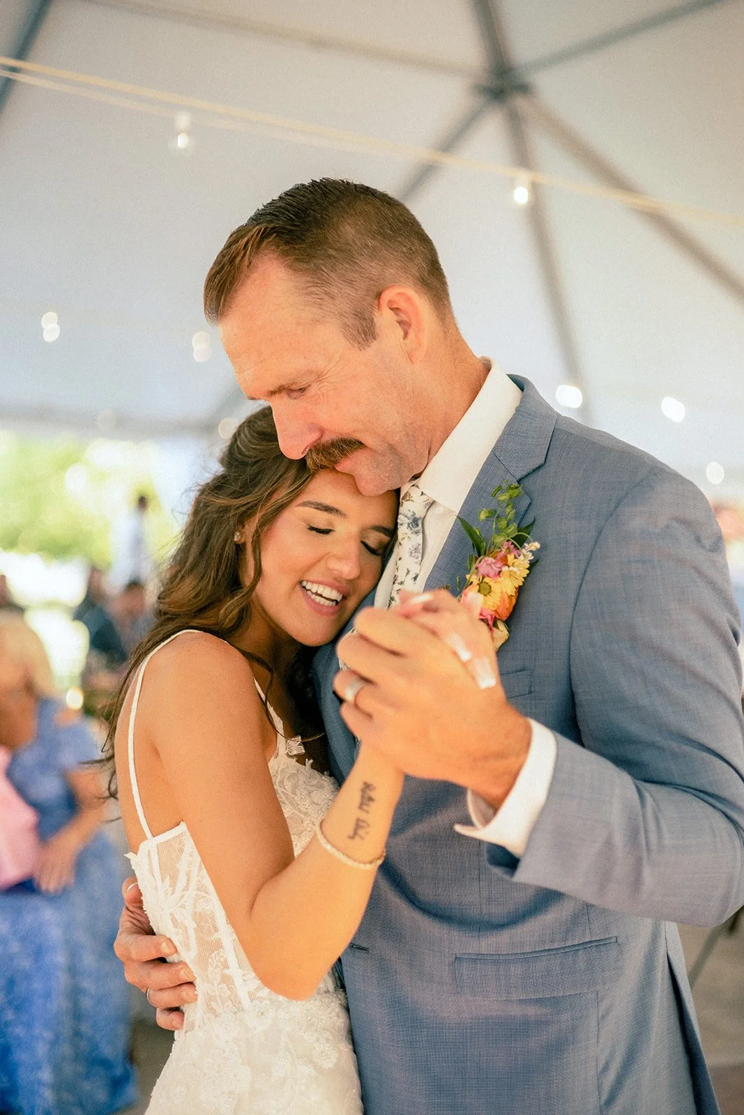 A bride and groom sharing a dance at their wedding, smiling with their eyes closed under a tent with string lights.
