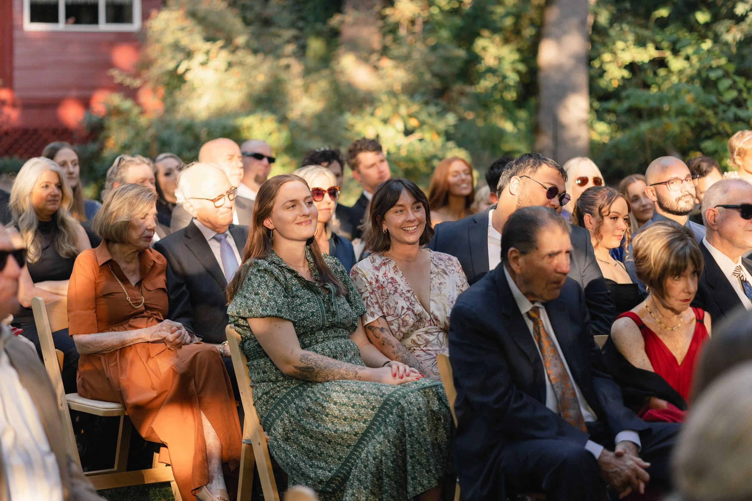 A diverse group of people sitting outdoors at an event, with some smiling and others focused, surrounded by trees and greenery in the background.