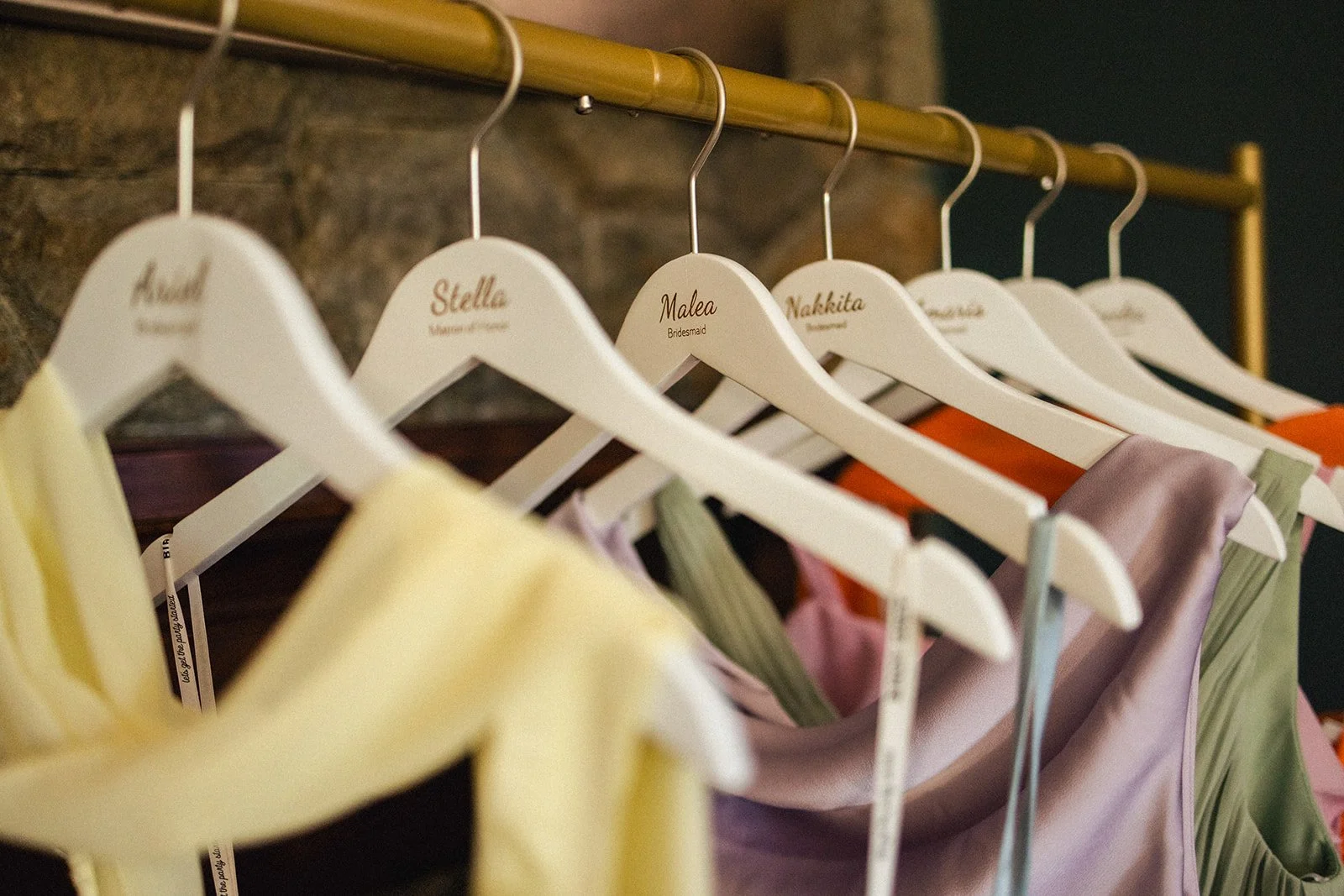 Colorful dresses hanging on a clothing rack with white velvet hangers labeled with names like Stella, Malea, Nakkita, and others, in a room with a stone or brick wall background.
