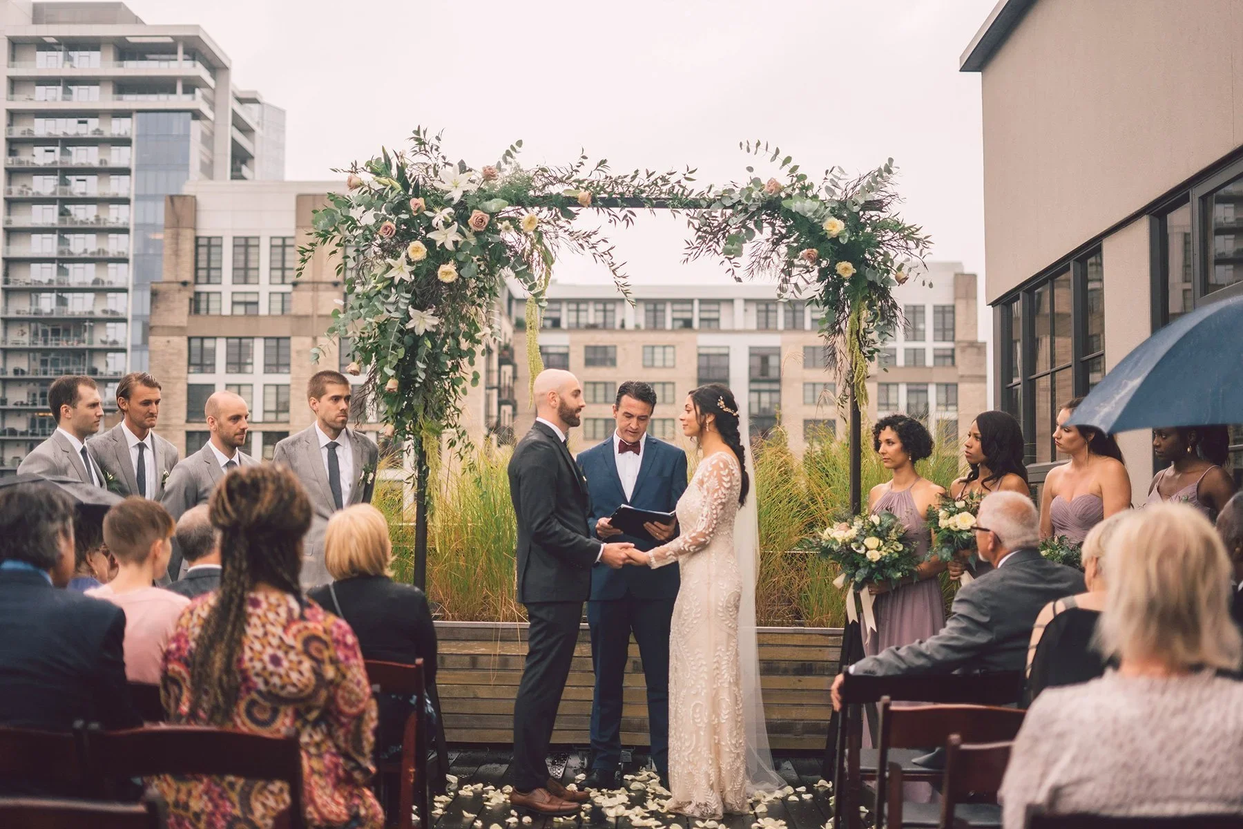 A wedding ceremony taking place outdoors on a rooftop, with the bride and groom exchanging vows under a floral arch, surrounded by friends and family.