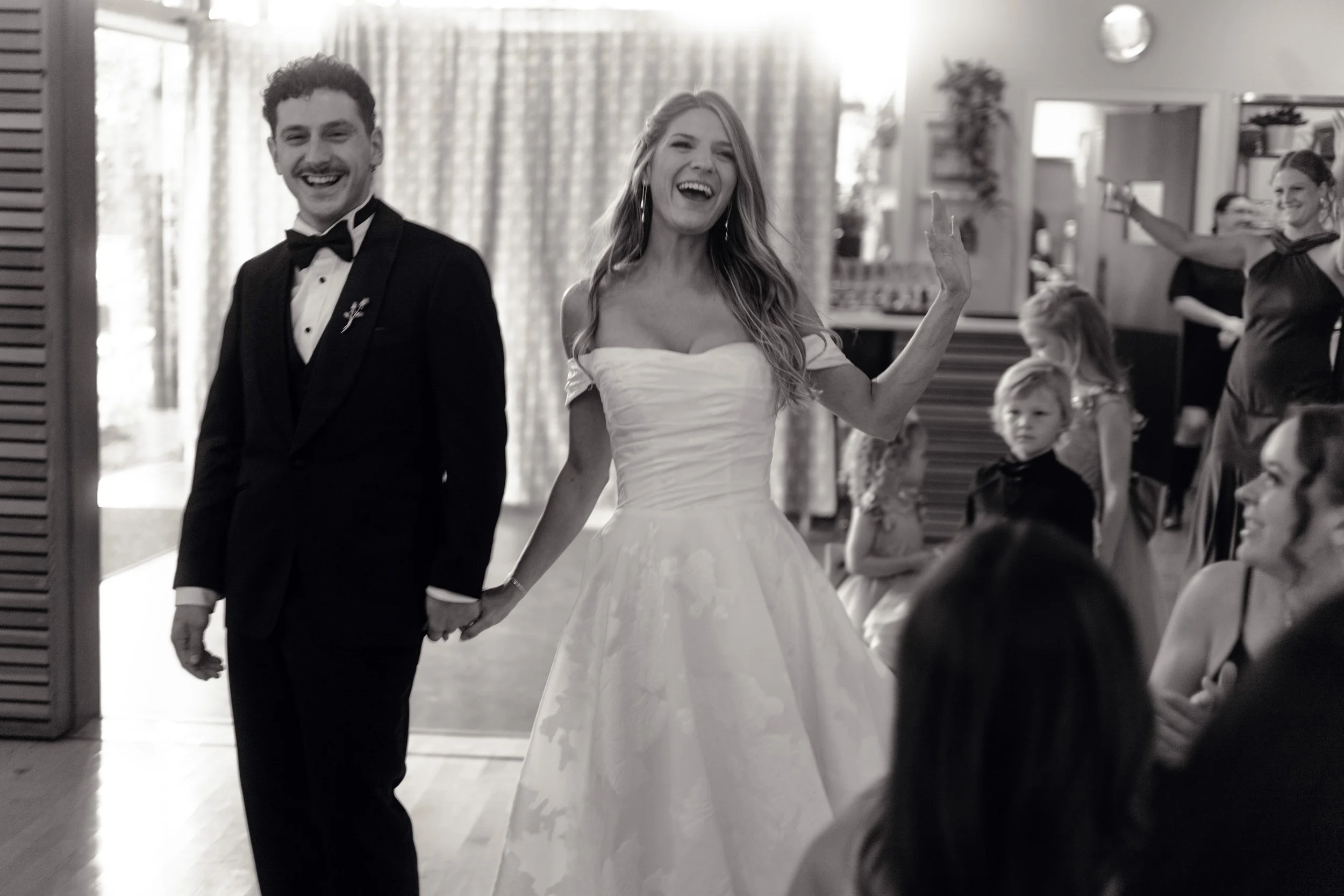A black-and-white photo of a bride and groom at their wedding reception, holding hands and smiling. The bride is wearing a white gown and the groom is in a black tuxedo, with people and children in the background.