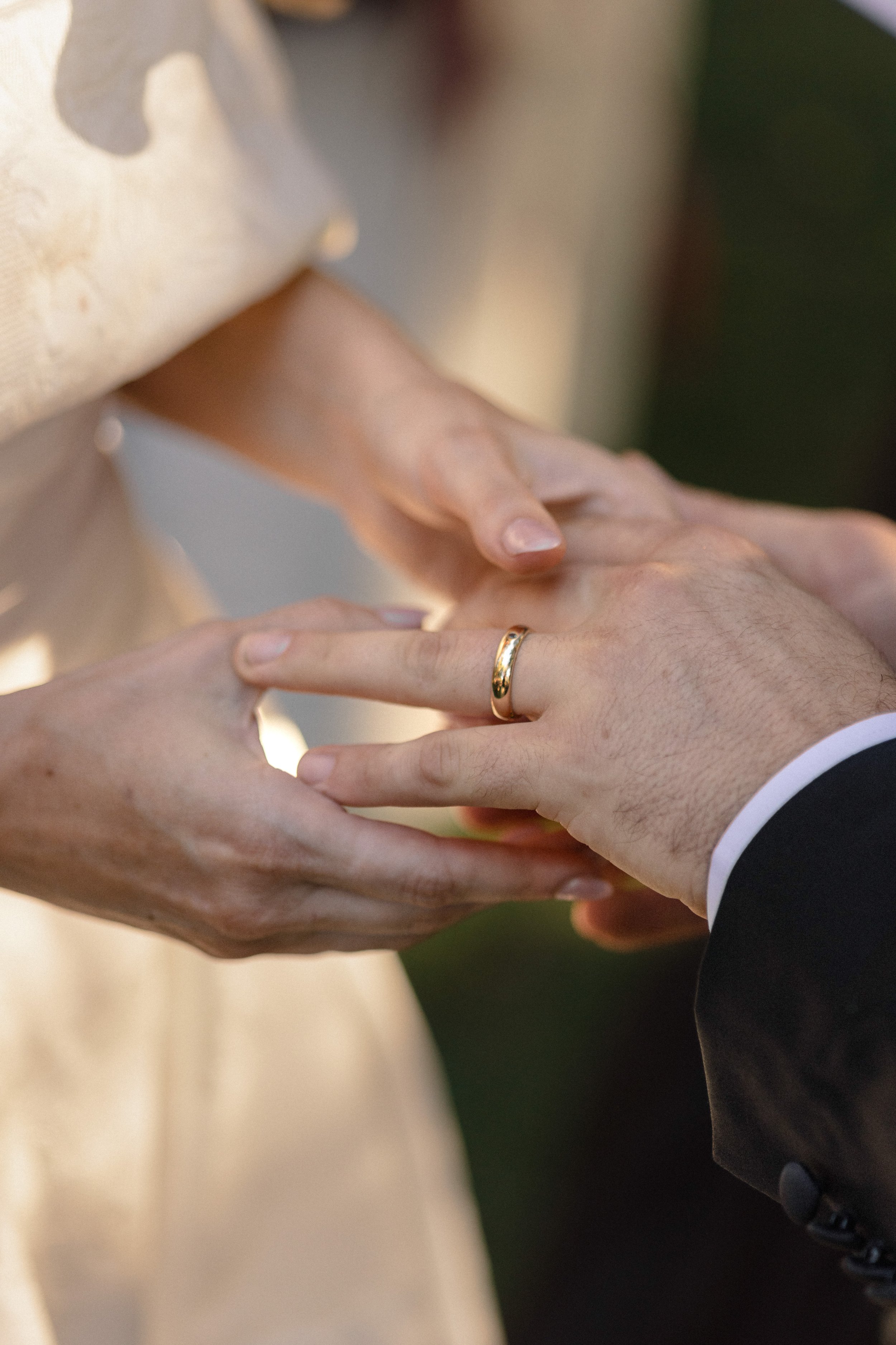 Close-up of a couple placing their hands together during a wedding ceremony, with the bride wearing a wedding dress and the groom in a black suit, both showing wedding rings.