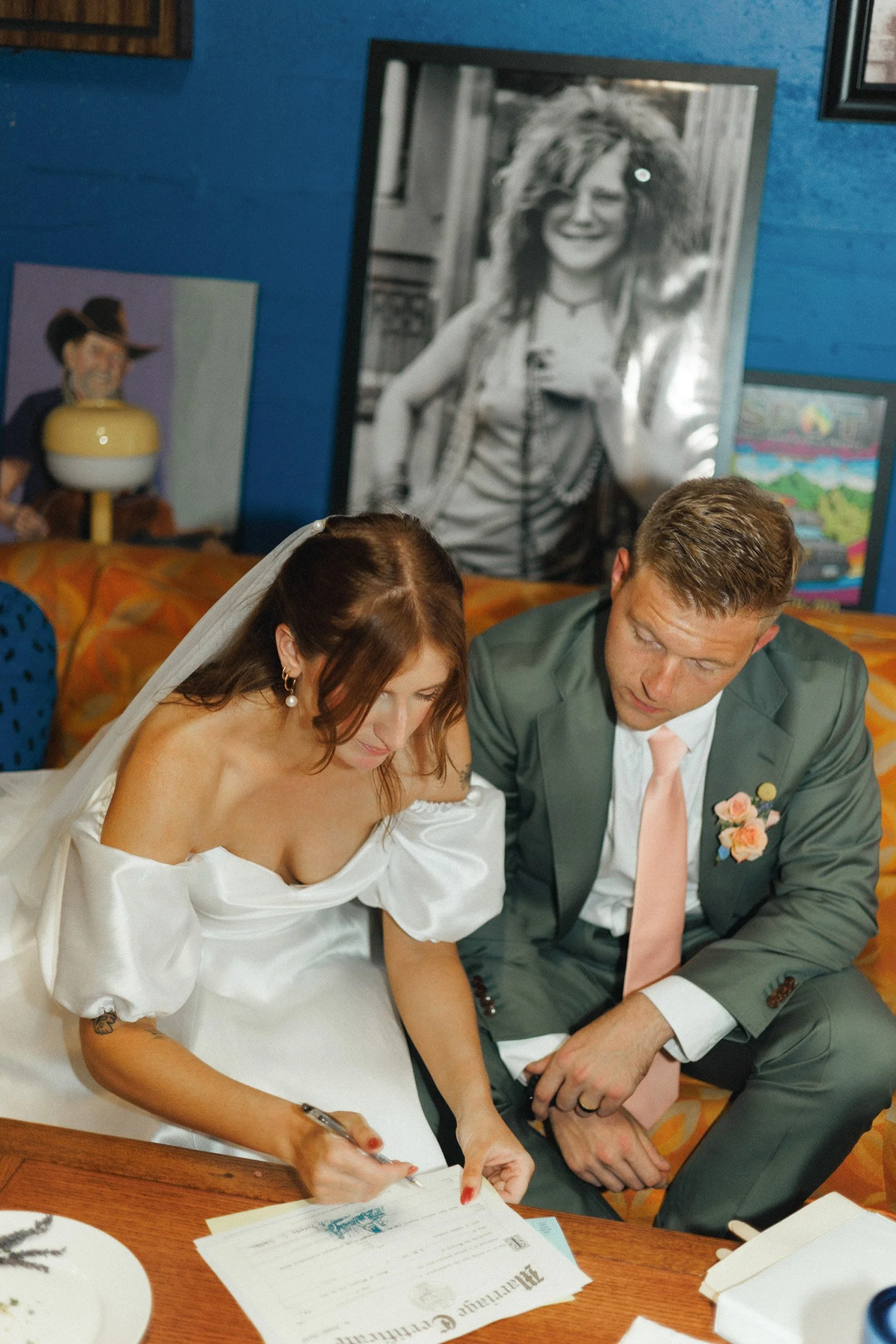 A bride in a white dress signs a document while a groom in a green suit watches, seated at a table in a colorful, art-filled room.