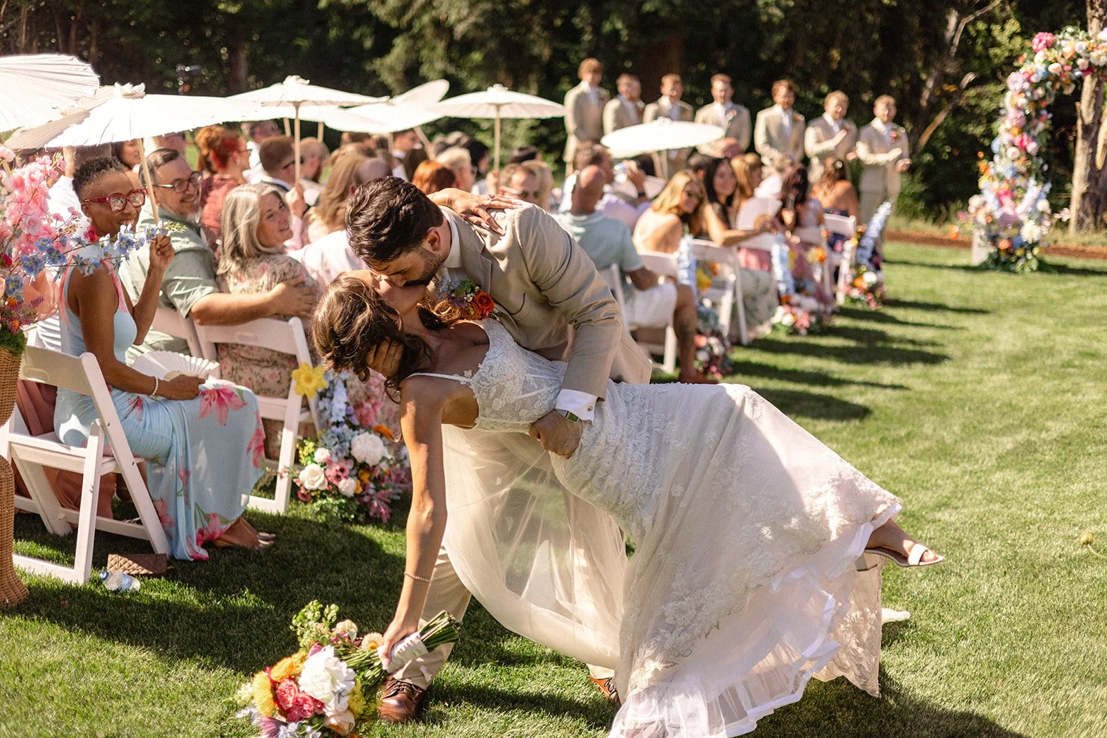 A newlywed couple sharing a kiss during their outdoor wedding ceremony as the groom dips the bride on the grass. Guests are seated with white umbrellas and floral decorations in the background.