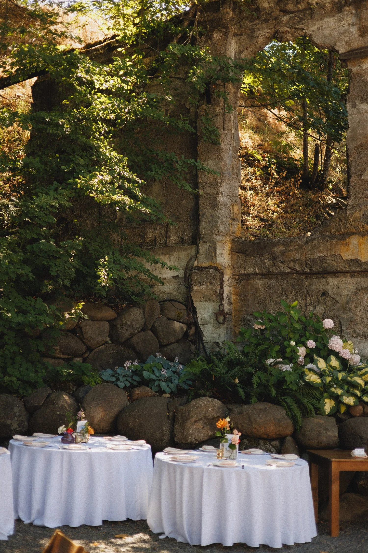Outdoor wedding reception with three tables covered in white cloth, decorated with colorful flower arrangements, set against an old stone wall and lush greenery.