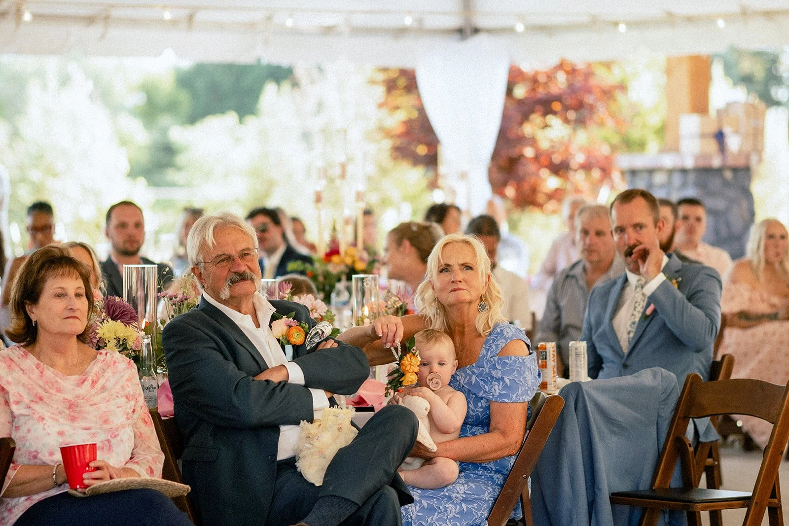 People gathered at a social event, seated at tables decorated with flowers, with a focus on an elderly man, woman, and a baby.
