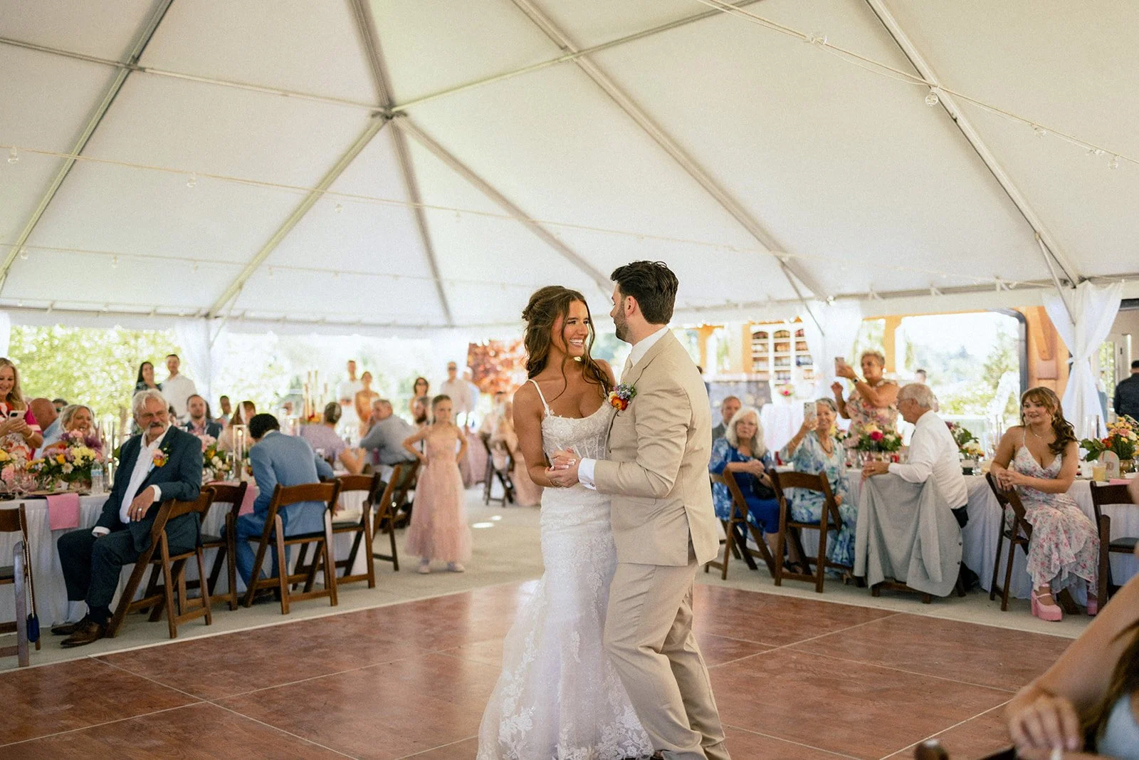 A bride and groom sharing their first dance at a wedding reception inside a tent, with guests seated at decorated tables watching and taking photos.