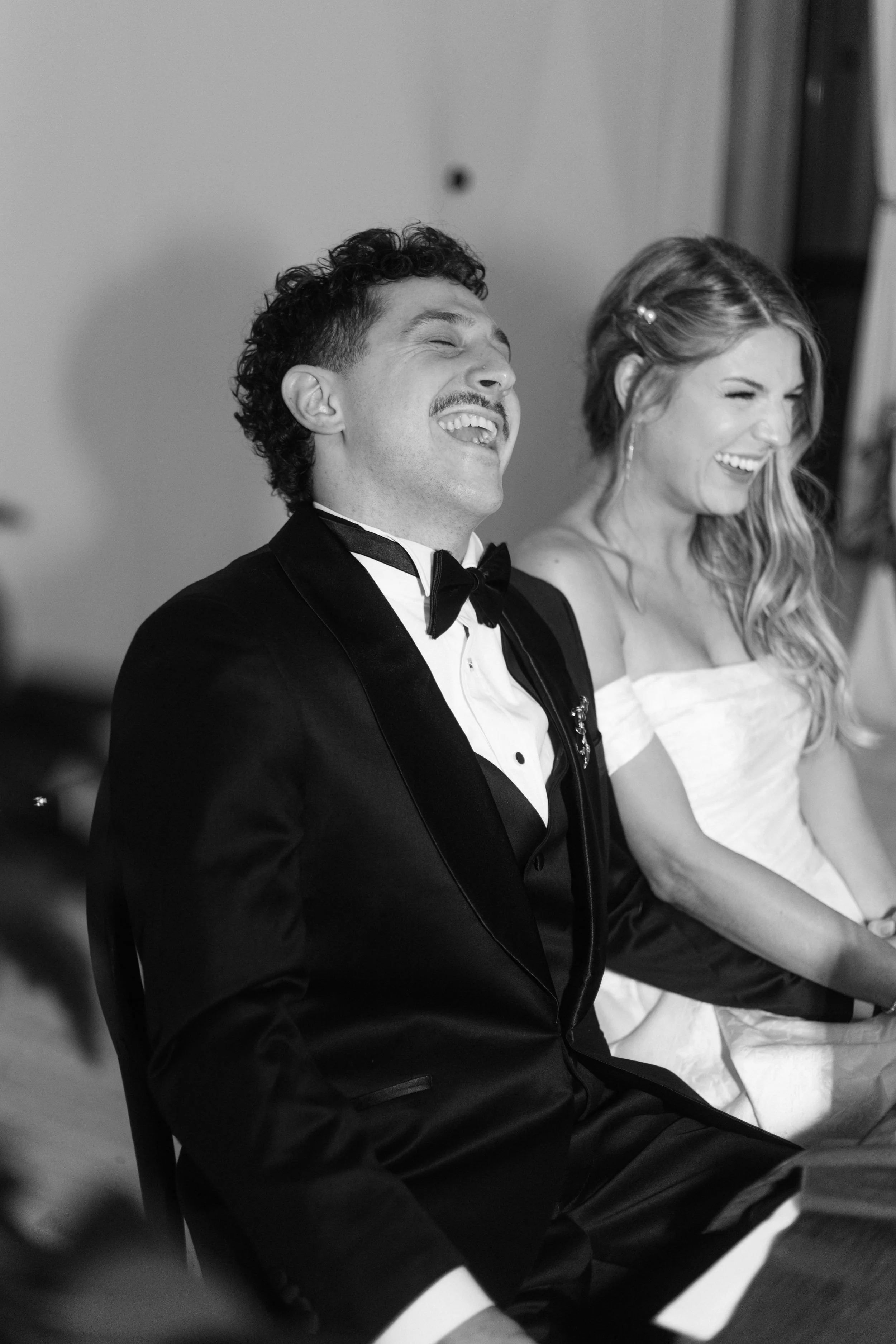 Black and white photo of a man in a tuxedo and woman in a white dress, both laughing and sitting close together.