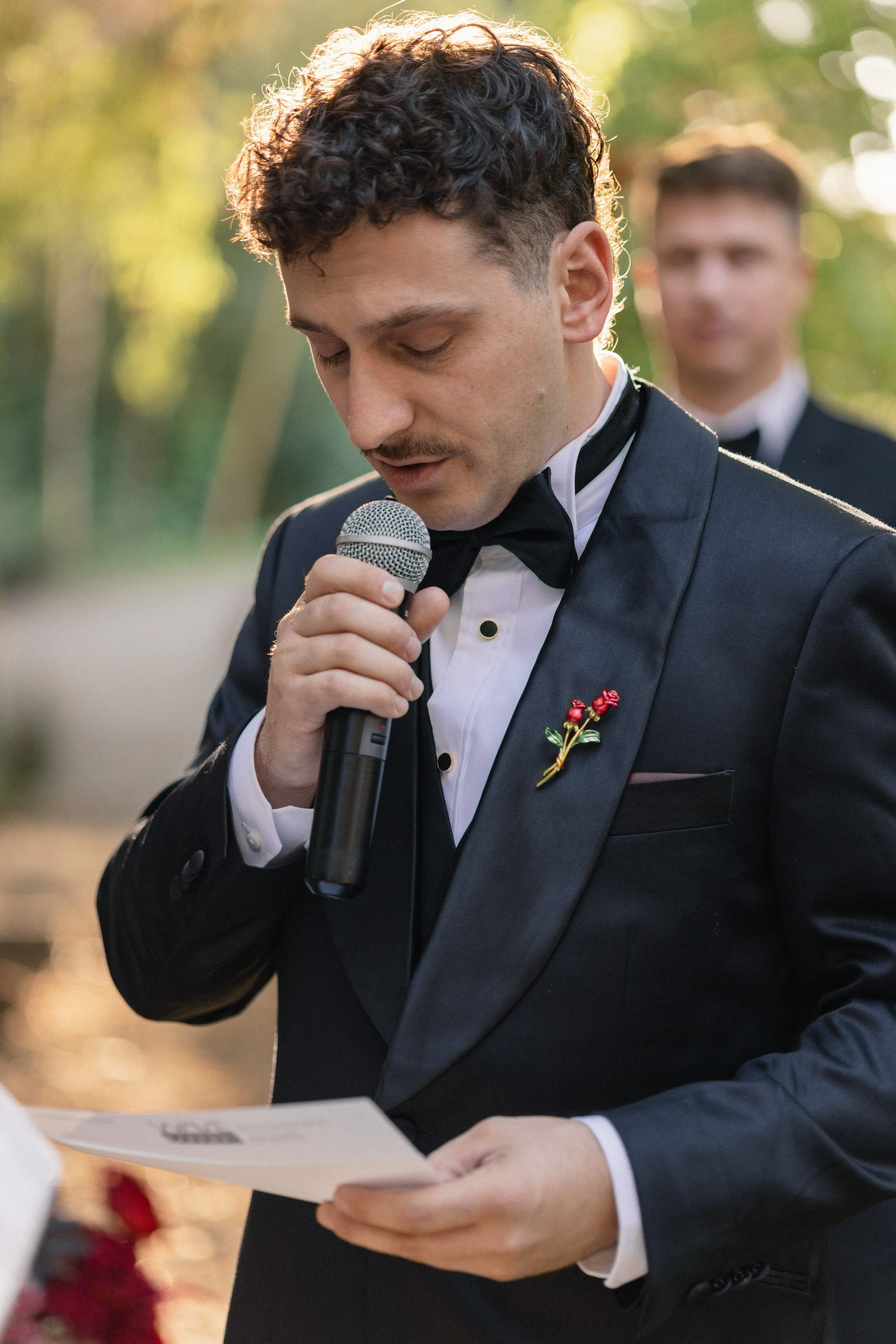A man in a tuxedo reading a speech into a microphone during an outdoor wedding ceremony, with a man in a tuxedo standing behind him, and blurred greenery in the background.