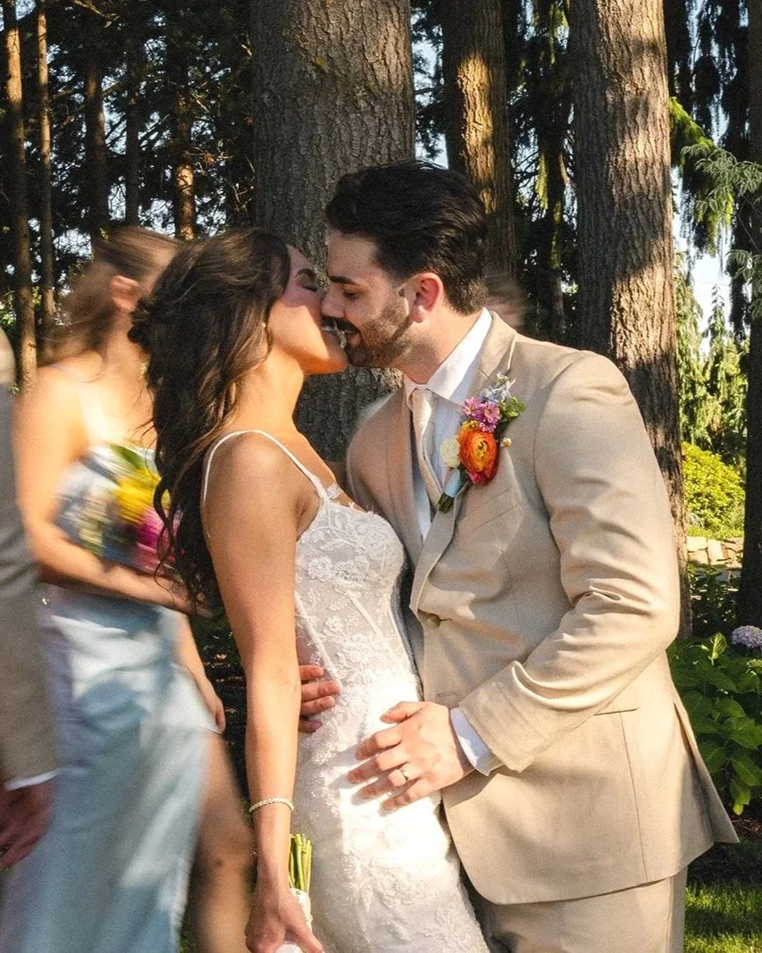 A bride and groom share a kiss on their wedding day outdoors in front of tall trees, with guests passing by in the background.
