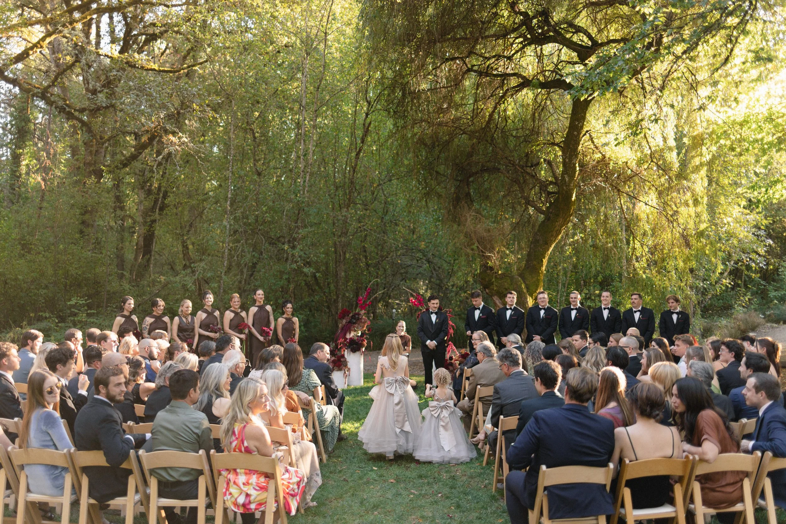 A large outdoor wedding ceremony with guests seated on wooden chairs, a bride and groom standing at the altar, surrounded by bridesmaids and groomsmen, in a wooded area with sunlight filtering through trees.