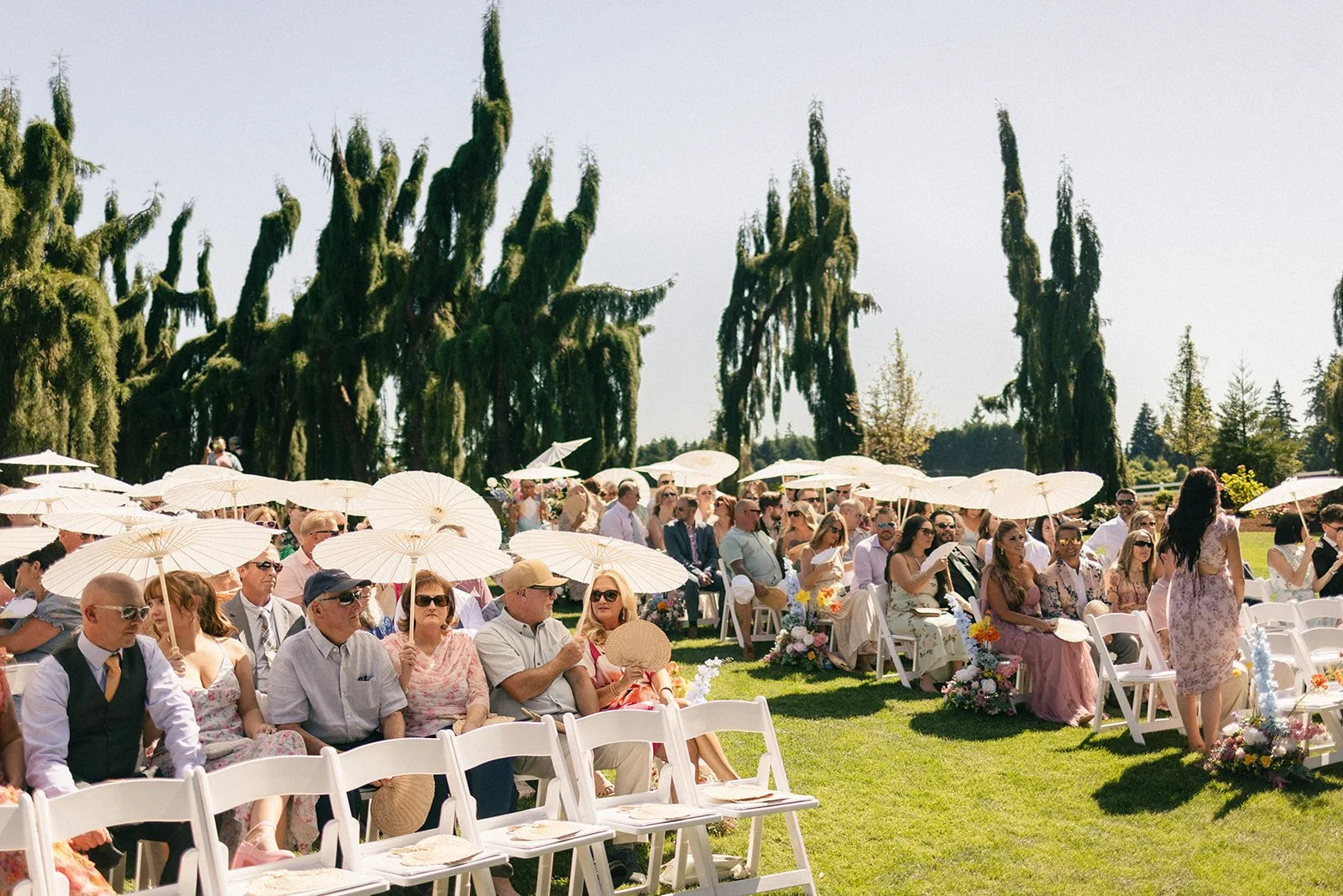 Guests seated outdoors at a wedding ceremony, holding parasols. The setting features tall trees and a bright, sunny sky.