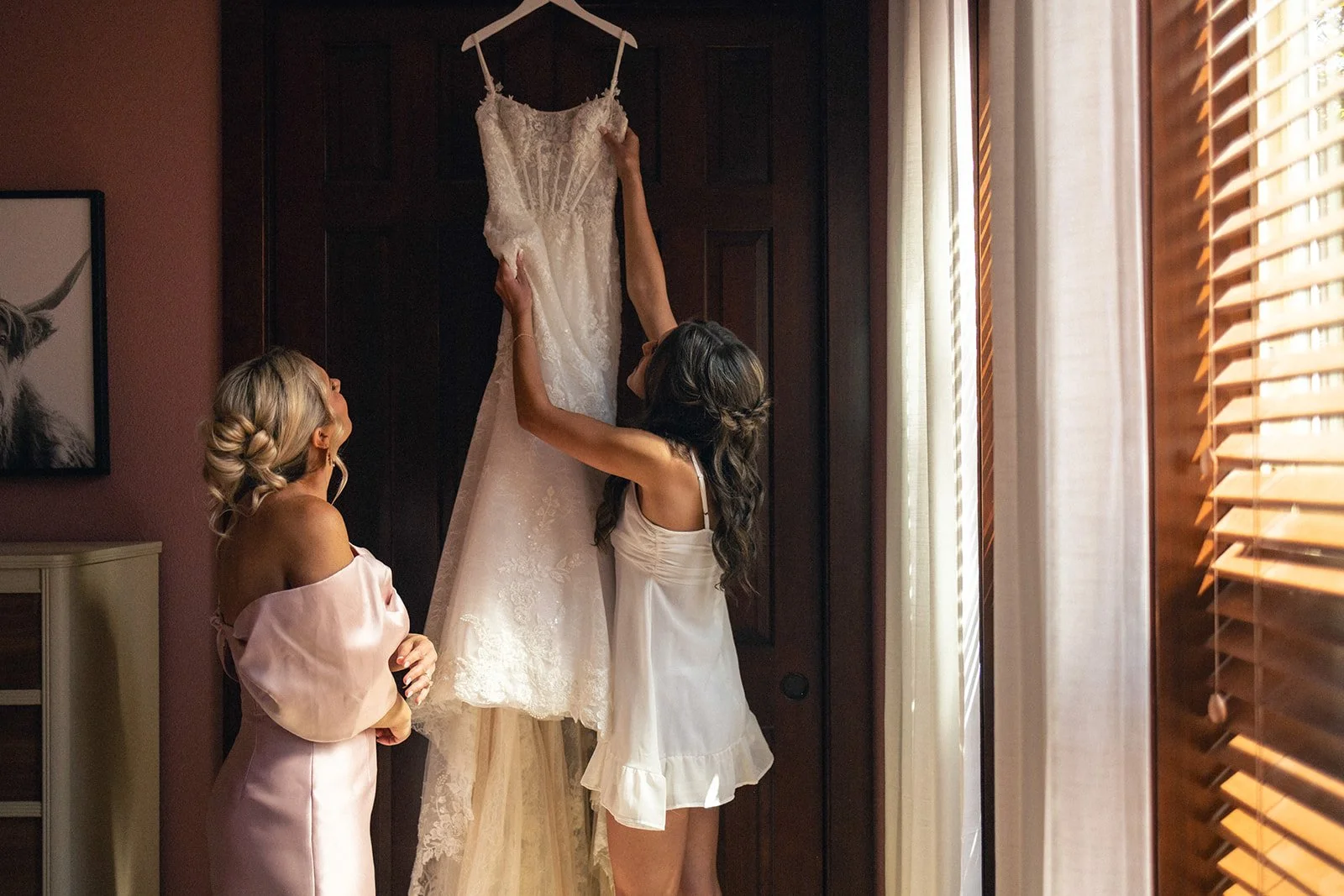 Two women, one with blonde hair in an updo and the other with long brown hair, are hanging a wedding dress on a dark wooden door. The room has natural light coming through window blinds on the right side.