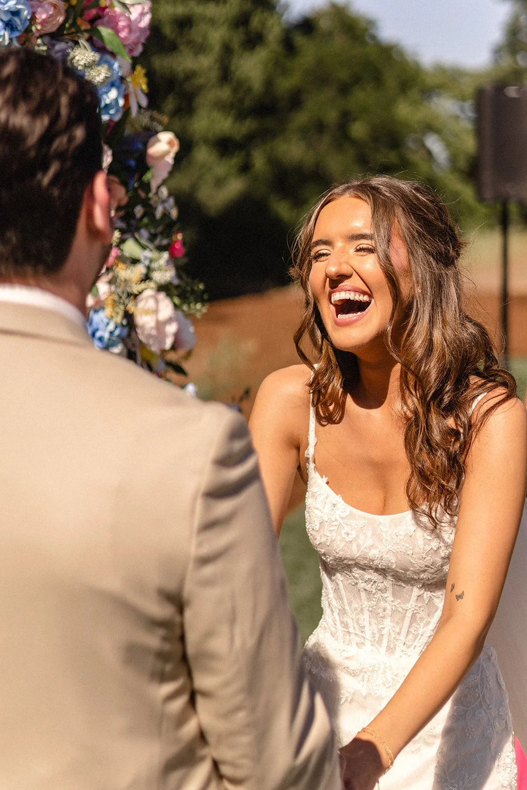 A woman in a white wedding dress laughing and standing outdoors during her wedding ceremony, with a man in a beige suit facing her, and a floral arch in the background.