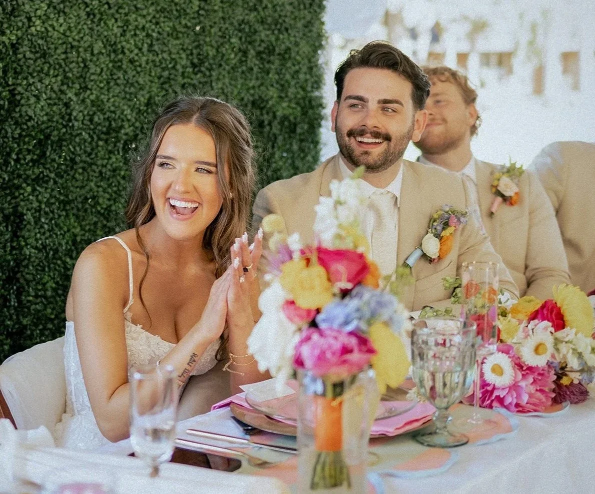 A bride and groom, both smiling, sitting at a decorated wedding reception table with flowers and glasses, enjoying the celebration.