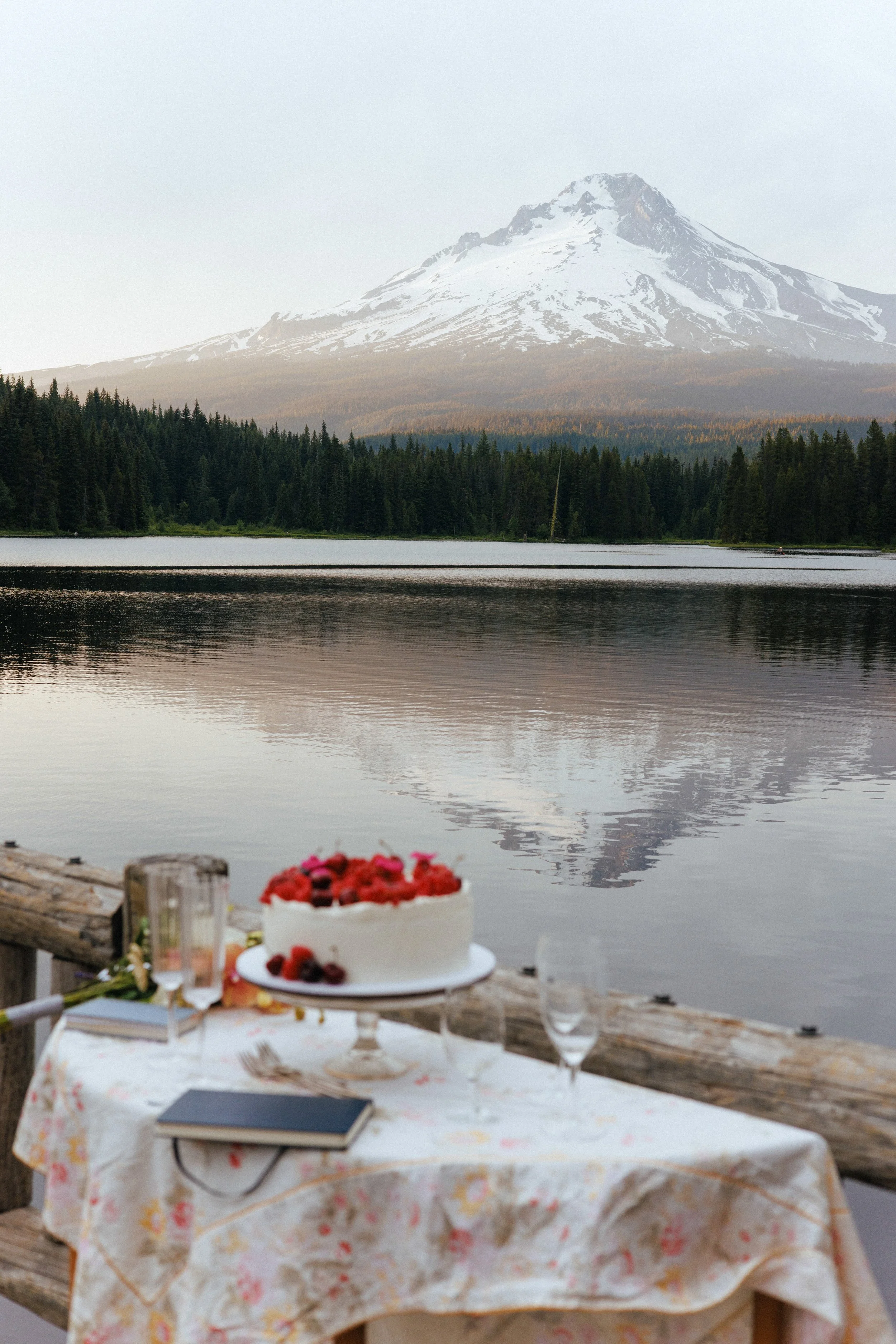 A lakeside scene with a mountain in the background, a wooden table with a floral tablecloth, and a birthday cake with berries on it in the foreground.