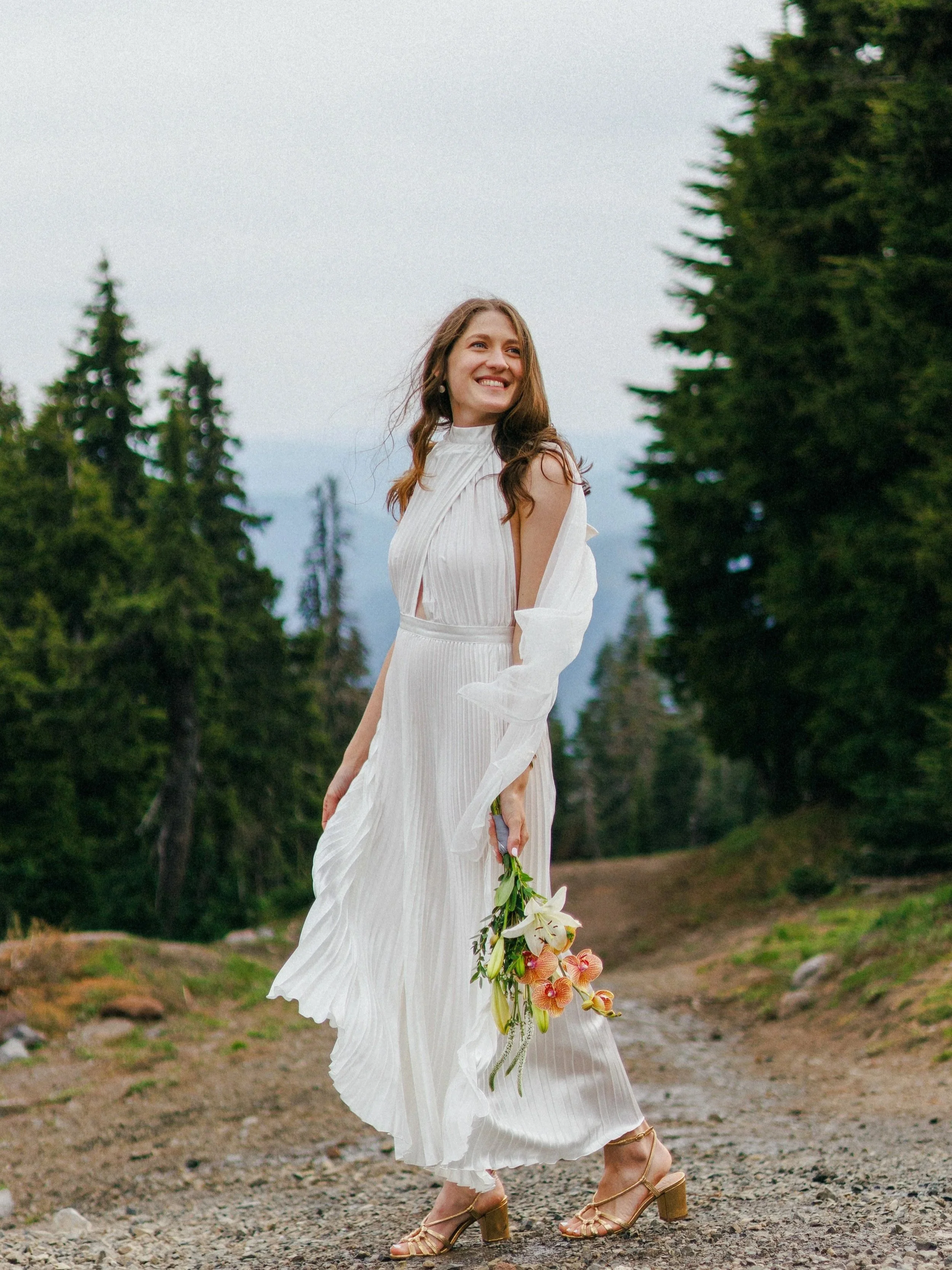 A woman in a white pleated dress holding a bouquet of flowers, standing outdoors on a dirt path surrounded by tall green trees, smiling and looking to the side.