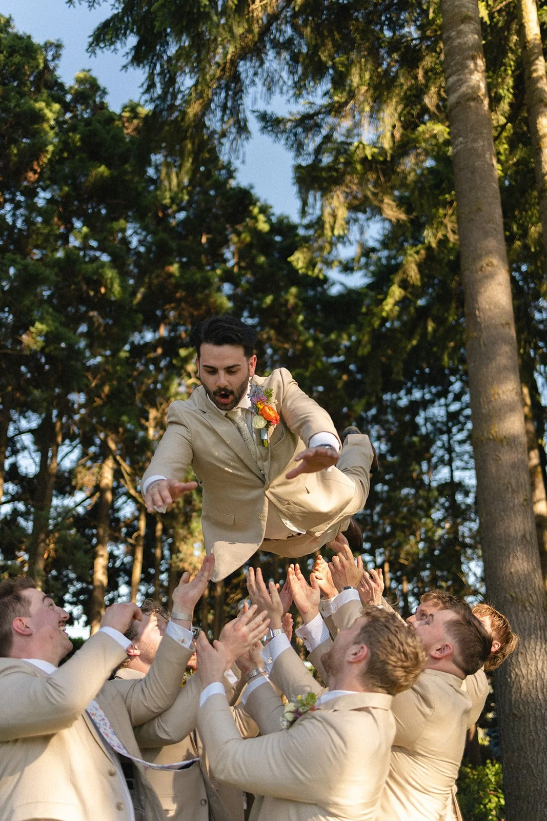 A man in a beige suit being thrown into the air by a group of men in matching suits outdoors with tall trees in the background.